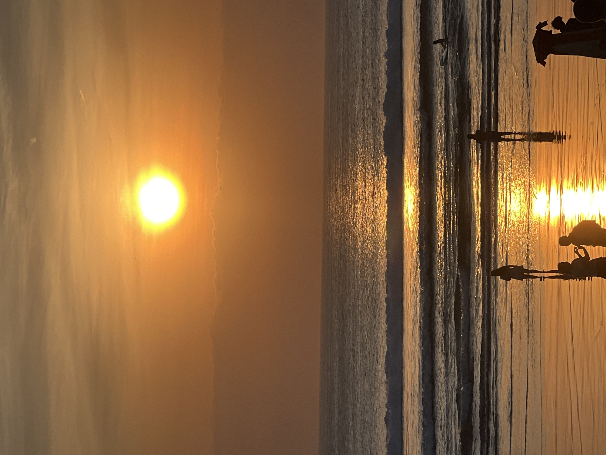 A vertical, medium shot captures a vibrant sunset over Kuta Beach, Indonesia. The sun, a bright orb of yellow and white, dominates the upper portion of the frame, casting a warm orange glow across the sky and reflecting brilliantly on the water's surface. Wispy clouds are visible near the horizon.
The ocean is a deep blue, with gentle waves rolling towards the shore, each crest highlighted by the setting sun's light. Several people are present on the wet sand and in the shallow water. In the foreground, silhouetted figures stand on the beach, their reflections shimmering on the wet sand. One figure stands near the water's edge, while two others are closer to the viewer, one appearing to take a photograph. Further out in the water, a person is standing and another is on a surfboard. The wet sand is smooth and appears highly reflective, mirroring the colors of the sunset. The overall atmosphere is serene and tranquil, typical of a sunset on this popular beach.