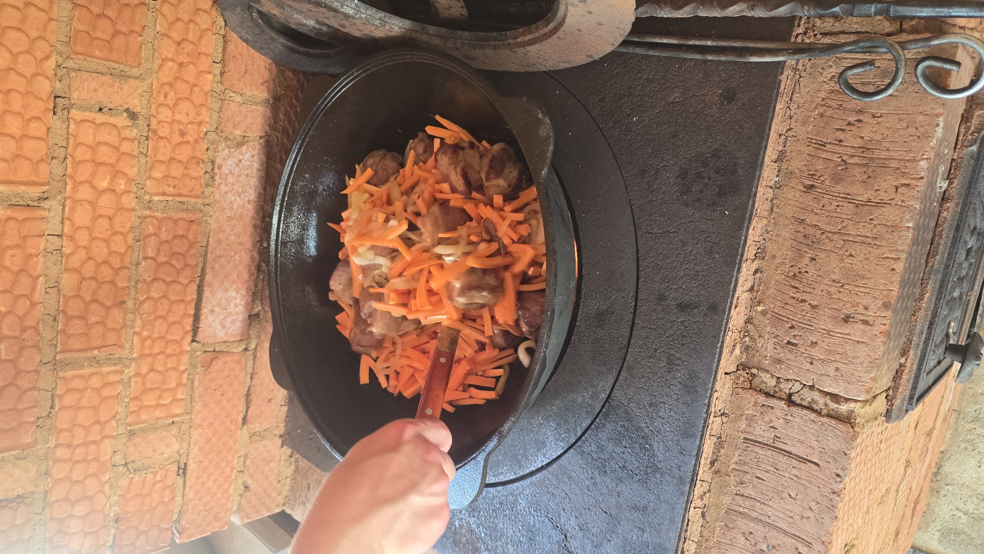 This image captures a cooking scene in Забегалово, Russia, featuring a large black cast-iron pot (kazan) resting on a dark, flat, possibly cast-iron stovetop. Inside the pot, browned chunks of meat are mixed with julienned orange carrots and white sliced onions. A light-skinned hand, entering from the left, uses a cooking utensil with a dark brown, possibly wooden, handle to stir the contents of the pot. The stovetop is part of a rustic brick structure. The background consists of a textured brick wall in shades of orange and red, with visible mortar. The bricks display a distinct, somewhat irregular, patterned surface. On the lower right, a dark, vertical element with decorative metal scrolls is partially visible. The lower section of the stove structure is also visible, made of similar brickwork, with a dark metal door or plate on the front.