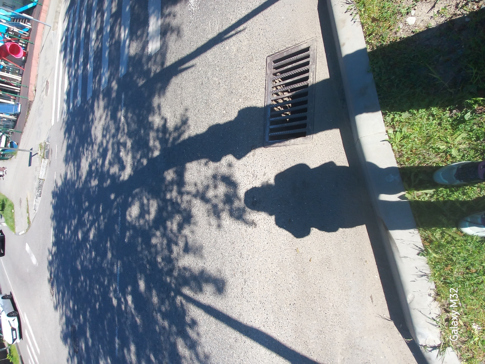 The image shows a street scene in Sochi, Russia, on a sunny day. Shadows of trees are cast across the asphalt, along with the shadow of a person. The street has a crosswalk, a storm drain, and a curb with grass growing alongside it. Several cars are parked along the edge of the street. There is playground equipment partially visible in the background. A phone logo "Galaxy M32" is in the corner of the image.