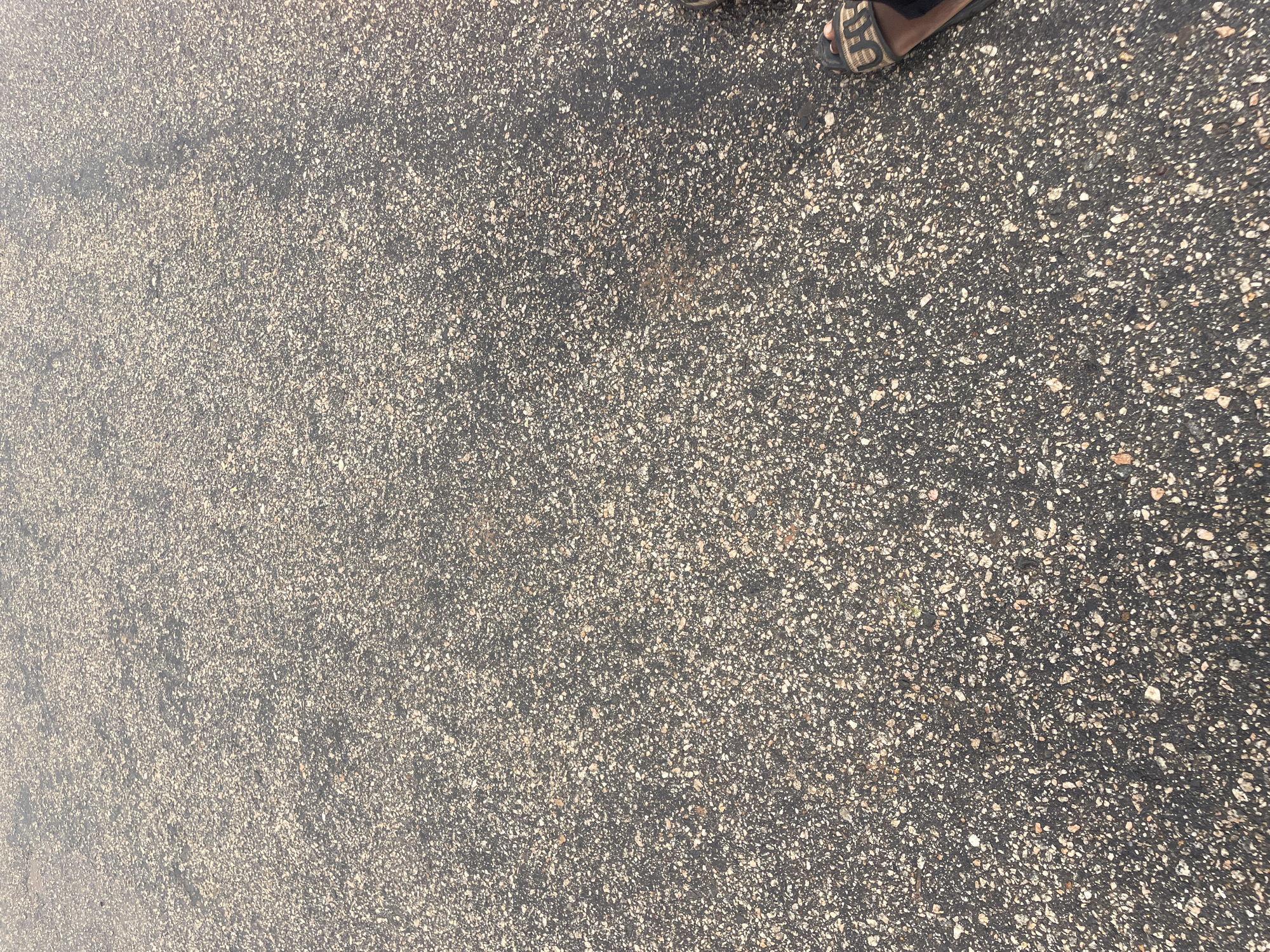 The image shows a close-up view of asphalt pavement. The surface is dark grey with numerous small, light-colored aggregate pieces embedded within it, giving it a speckled appearance. The texture is rough and uneven. In the bottom right corner of the image, a person's foot wearing a dark sandal with a woven pattern is visible. The sandal has a strap that goes over the toes and a heel strap. The scene appears to be outdoors, likely on a street or walkway in the city of Lokoja, Nigeria, given the context provided. There is no other identifiable activity or interaction occurring in the scene.