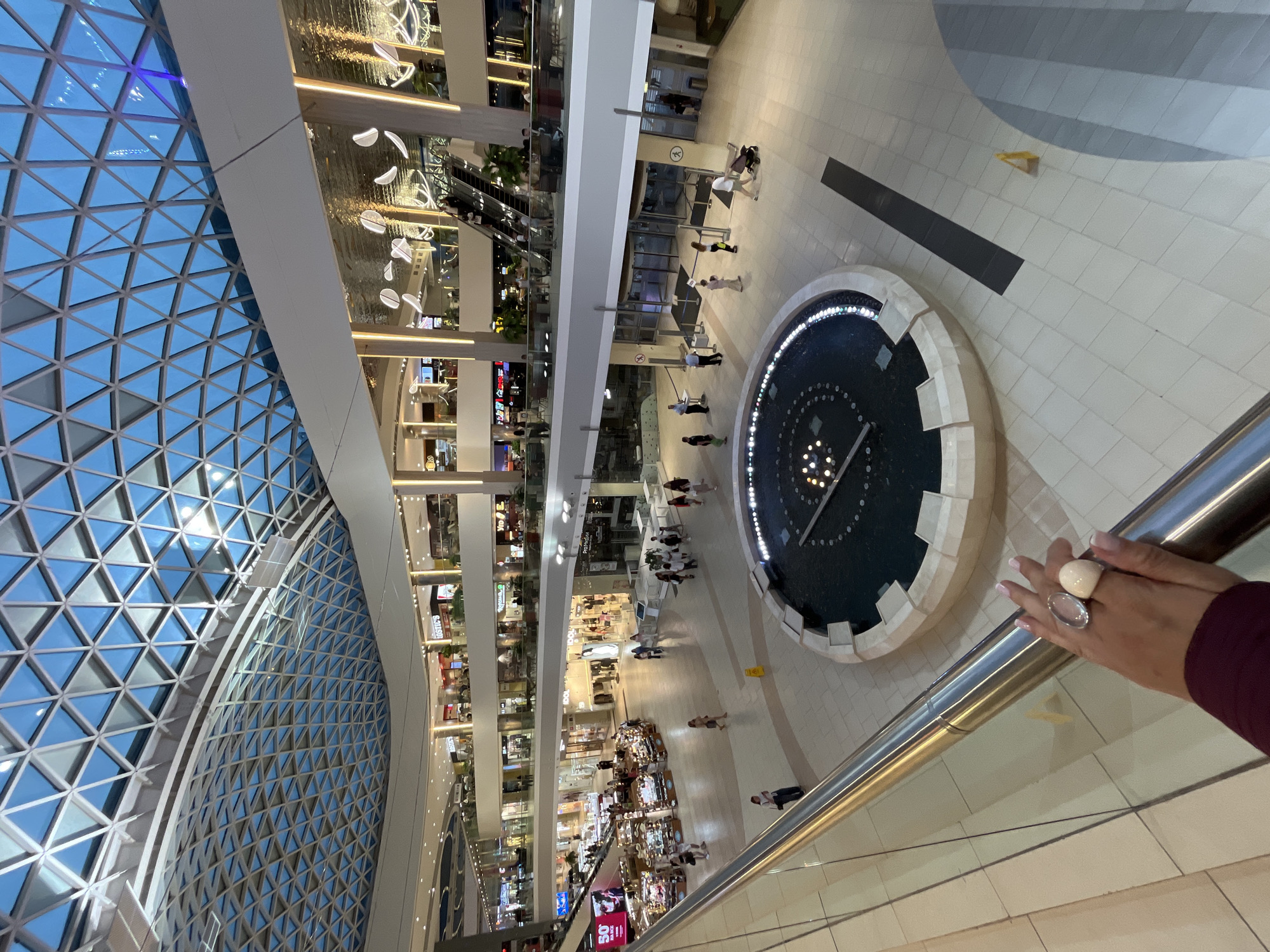 The image shows an interior view of a modern shopping mall, likely in Sochi, Russia, characterized by its multi-level design, extensive glass ceiling with a geometric pattern, and contemporary lighting fixtures. In the foreground, a hand with rings and manicured nails rests on a silver railing, suggesting the photographer's perspective. The mall's interior is brightly lit, with various shops and escalators visible on different levels. People are dispersed throughout the space, some walking, others standing or sitting, indicative of a typical commercial environment. A prominent feature in the central area is a circular fountain with lights in the water. The overall impression is one of a well-maintained and bustling shopping complex.