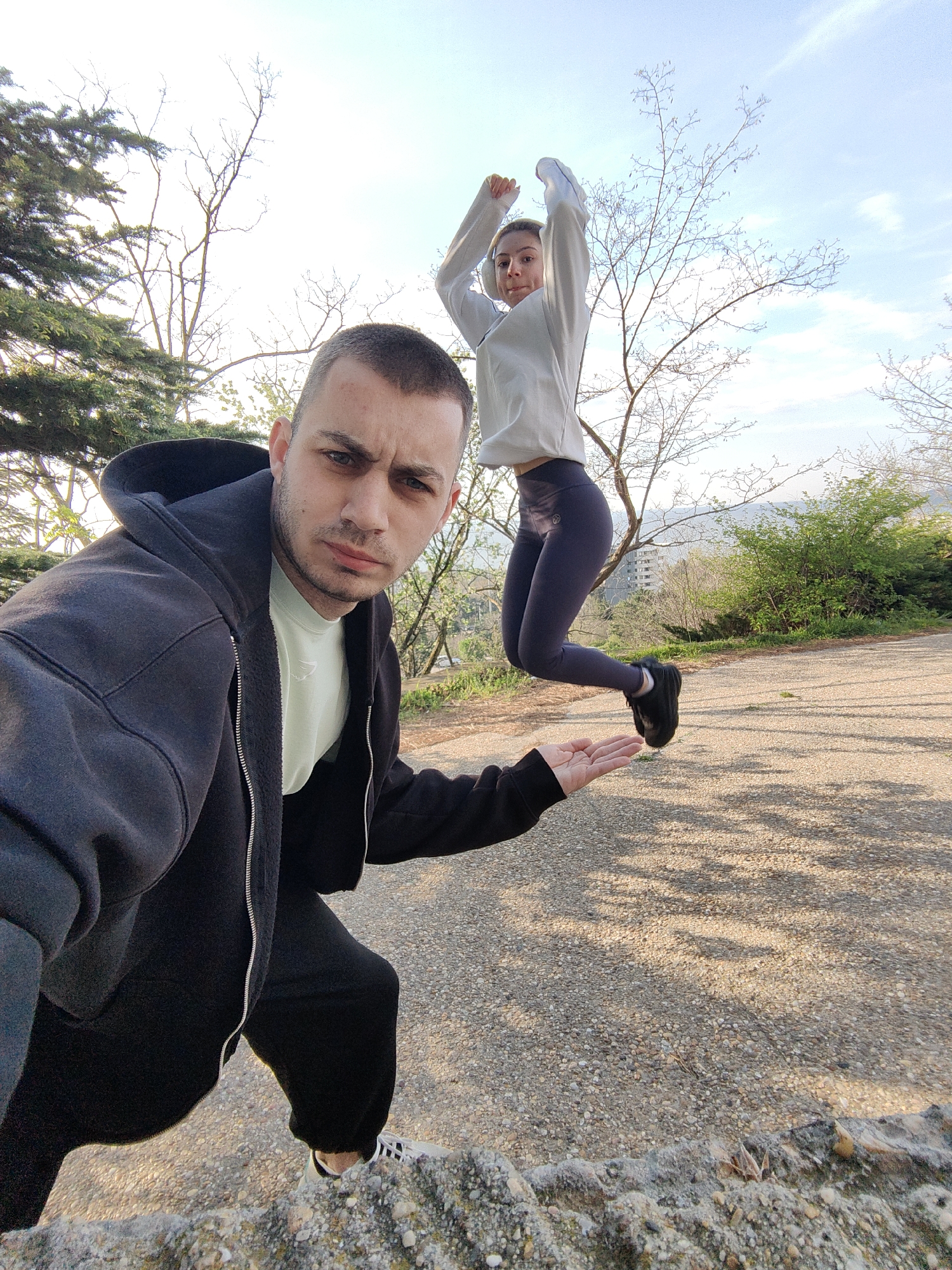 The image captures two individuals in an outdoor setting, likely a park in the city of Varna, Bulgaria. A man in the foreground is positioned near a low stone wall, facing the camera. He is wearing a black hooded jacket over a light-colored t-shirt and black pants. His stance suggests he is crouching or kneeling. To his right and further in the background, a woman is captured mid-jump. She has her arms raised, wearing a light-colored top, dark leggings, and black sneakers. The ground appears to be a paved pathway with scattered shadows and the natural landscape of trees and greenery provides a backdrop. The sky is partially cloudy.
