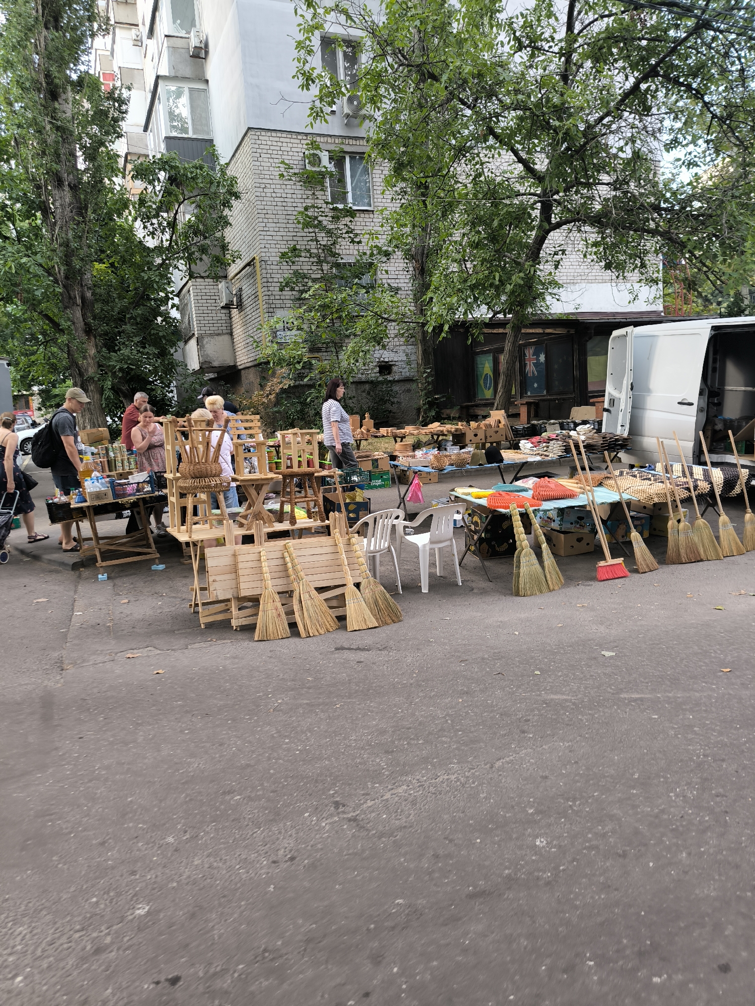 This image captures an outdoor market scene set against the backdrop of a multi-story residential building and trees. The market appears to be a makeshift setup, with tables and stands displaying various goods. Prominently featured are numerous brooms, some neatly arranged in rows, while others are leaning against display structures. Wooden furniture, including small tables and chairs, are also visible. Several people are browsing or attending to the stalls. In the background, a white van with its rear doors open is parked, and beyond that, parts of the brick building and trees create a sense of an urban environment. The ground is asphalt. This scene is consistent with an informal market or flea market that might be found in residential areas of Mykolaiv, Ukraine, offering goods like household items and handmade crafts.