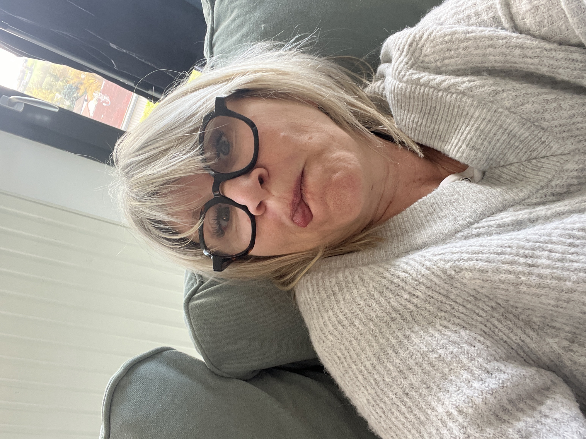 A medium shot depicts a fair-skinned, blonde woman with visible wrinkles and wearing black-rimmed glasses lying on a green upholstered chair. The woman is partially obscured by a light grey knit sweater. Her tongue is slightly protruded. In the background, a window reveals an external scene with foliage, indicating a location likely within Chambœuf, France.
