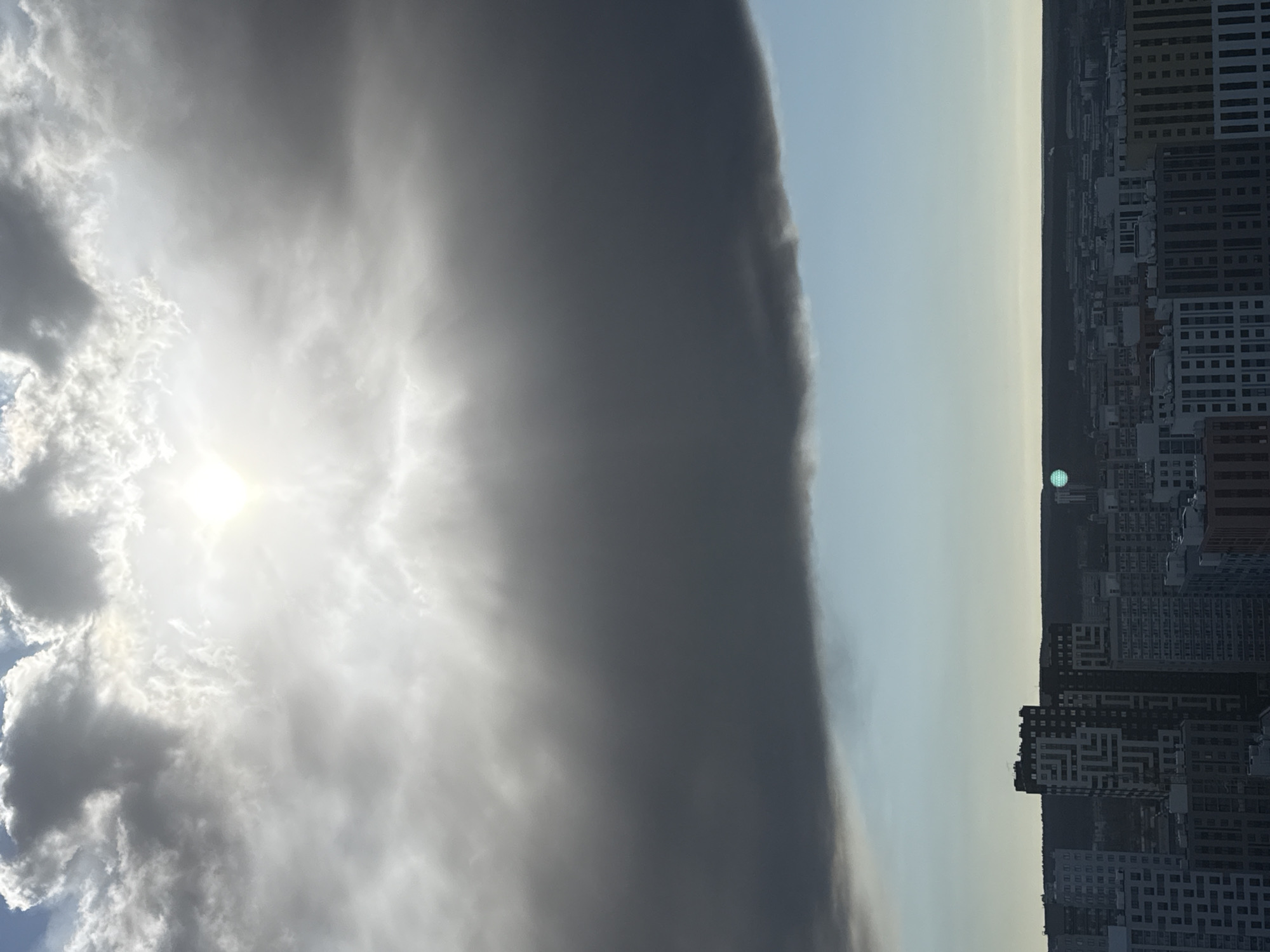 The image, rotated 90 degrees counter-clockwise for correct orientation, presents a dramatic panoramic view of the city of Perm, Russia, under a striking sky. The scene is bisected vertically by contrasting weather phenomena.
On the left, the sky is bright and partially clear, dominated by the sun shining intensely through a scattering of white and grey clouds, with visible rays emanating outwards. This area suggests fair or clearing weather.
To the right, a massive, dark grey cloud front stretches across the sky, forming an imposing, almost vertical wall that sharply divides the sky's light and dark halves. This prominent feature suggests an approaching storm or a significant weather system. Beyond this dark cloud, the sky becomes a lighter, hazy blue-grey towards the horizon.
Below this dramatic sky lies a dense urban landscape characterized by numerous multi-story buildings forming a bustling city skyline. Many of these structures appear dark, either silhouetted against the brighter sky or under the shadow of the storm front. In the far distance, a faint, dark line likely indicates a natural horizon of hills or forest beyond the city. A small, circular, greenish-blue object is visible in the distant, lighter sky, positioned near the horizon line above the cityscape.
The time of day is clearly daytime, with the bright sun suggesting late morning or afternoon. The overall impression is one of impending weather change or a powerful natural display over an urban environment. No people or visible text are present in the image.