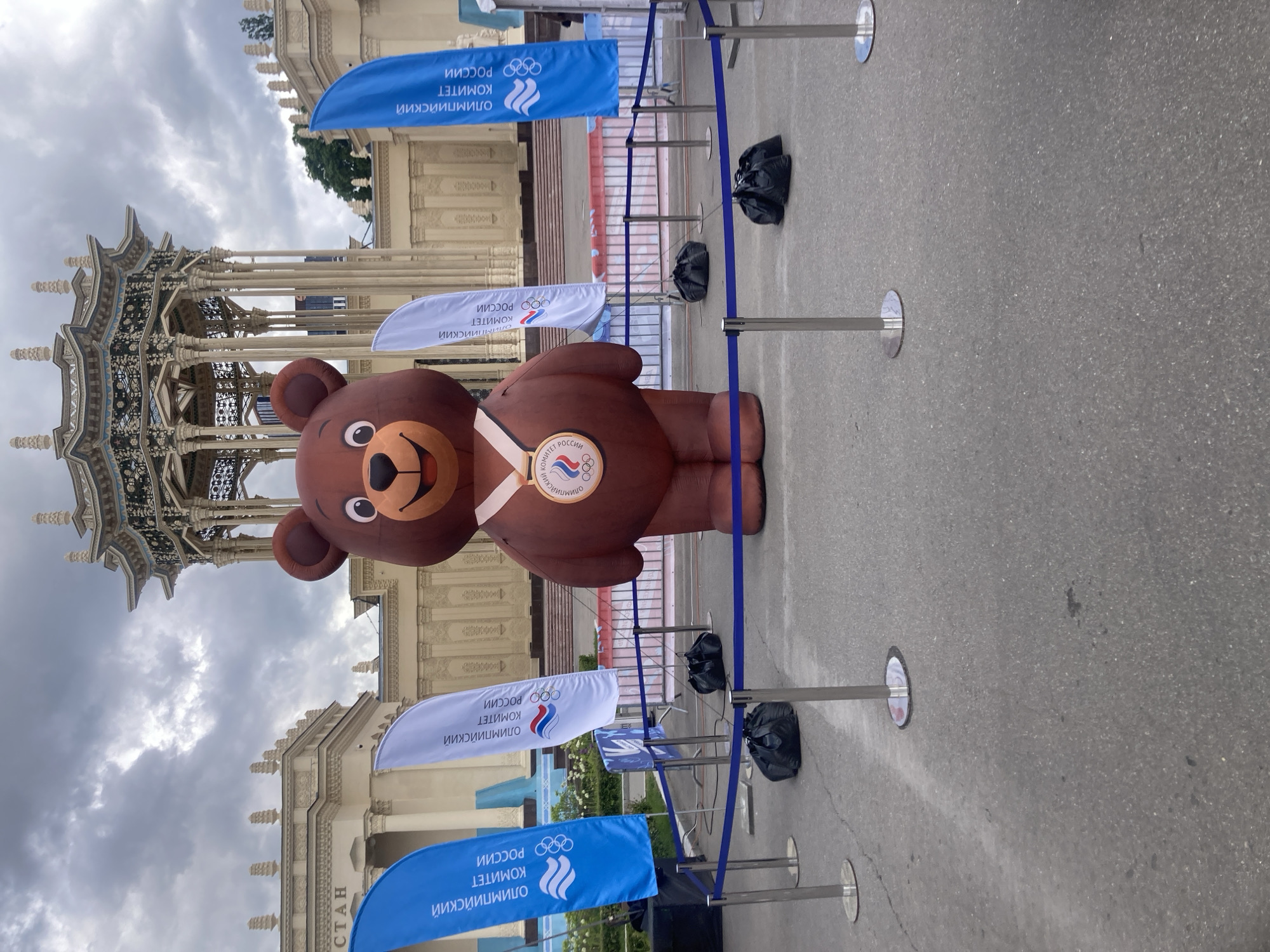 Here's a detailed description of the image:
**Overall Scene:** The image appears to be taken outdoors, possibly at an event or public space in Moscow, Russia. The weather is overcast.
**Main Subject:** A large inflatable brown bear with a cheerful face is positioned as a focal point. The bear wears a white sash/ribbon diagonally across its body, with a circular logo representing the Russian Olympic Committee.
**Surrounding Elements:**
* **Barricades:** The bear is surrounded by metal posts connected with blue tape/ribbon, forming a perimeter or barricade around the figure. There are black bags attached to the posts.
* **Flags/Banners:** Various flags or banners are placed around the scene. These flags are white and blue. The banners feature the Russian Olympic Committee emblem and text in Russian.
* **Building:** In the background, there is an ornate building or structure with intricate architectural details.
* **Ground Surface:** The ground is paved and appears to be concrete.
**Environment:** The environment is a public space. The presence of the inflatable bear and the Russian Olympic Committee branding suggest this is likely an event related to sports or the Olympics.