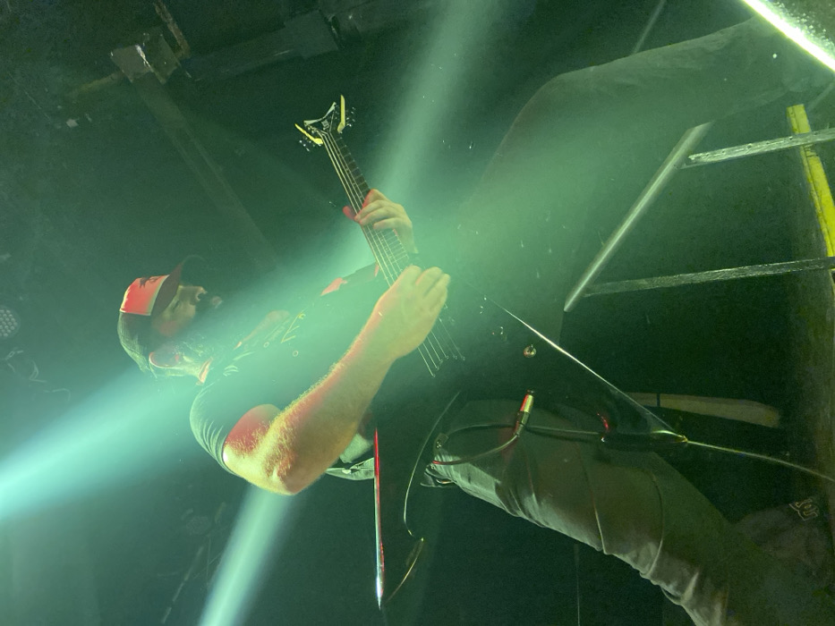 A man plays a black V-shaped electric guitar while positioned on a scaffolding platform. He wears a black t-shirt and dark pants, along with a red and black baseball cap. The stage is illuminated by a bright green spotlight, which creates a hazy effect. The angle is from below, looking up at the guitarist, with scaffolding visible in the background.