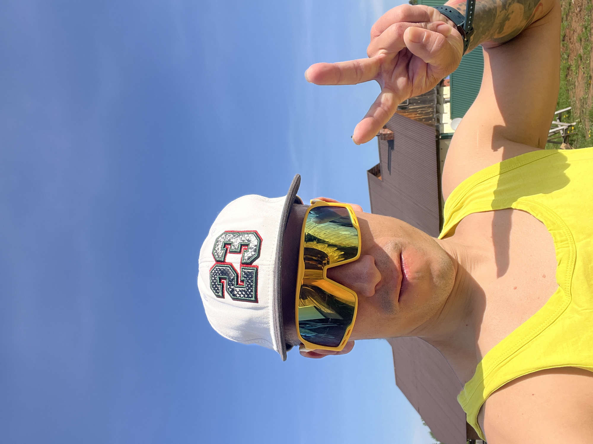 The image shows a man outdoors, likely in Chaikovsky, Russia, based on the potential location information. He is wearing a white baseball cap with a large "23" emblem, yellow-framed sunglasses with reflective lenses, and a yellow tank top. He is making a hand gesture with his index and middle fingers extended in a "peace" or "victory" sign. The background includes a clear blue sky, a brown wooden building, and green grass. The man appears to be the primary subject, positioned at the center of the frame.