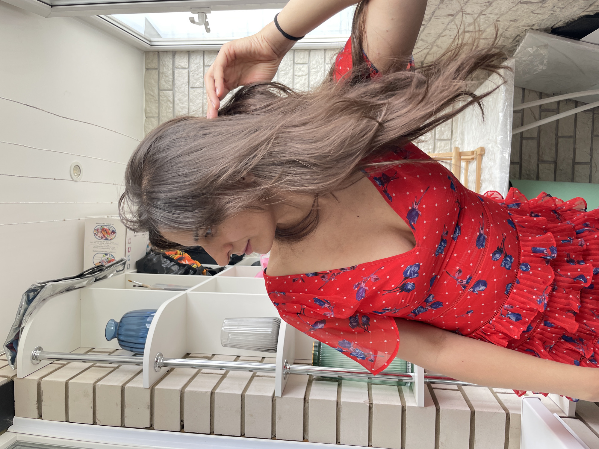 A woman with long, brown hair is posed indoors, looking to her left with her right hand touching her hair. She is wearing a red, short-sleeved dress with a floral pattern. The background consists of a white shelf unit with various decorative items, including a blue vase and a tall glass vase, positioned against a brick-patterned wall. A window is visible to the right, and the ceiling has visible cables and lighting. The scene appears to be a balcony or a room with access to an outdoor space, possibly located in Izhevsk, Russia, as indicated by the context. The overall lighting suggests a daytime setting.