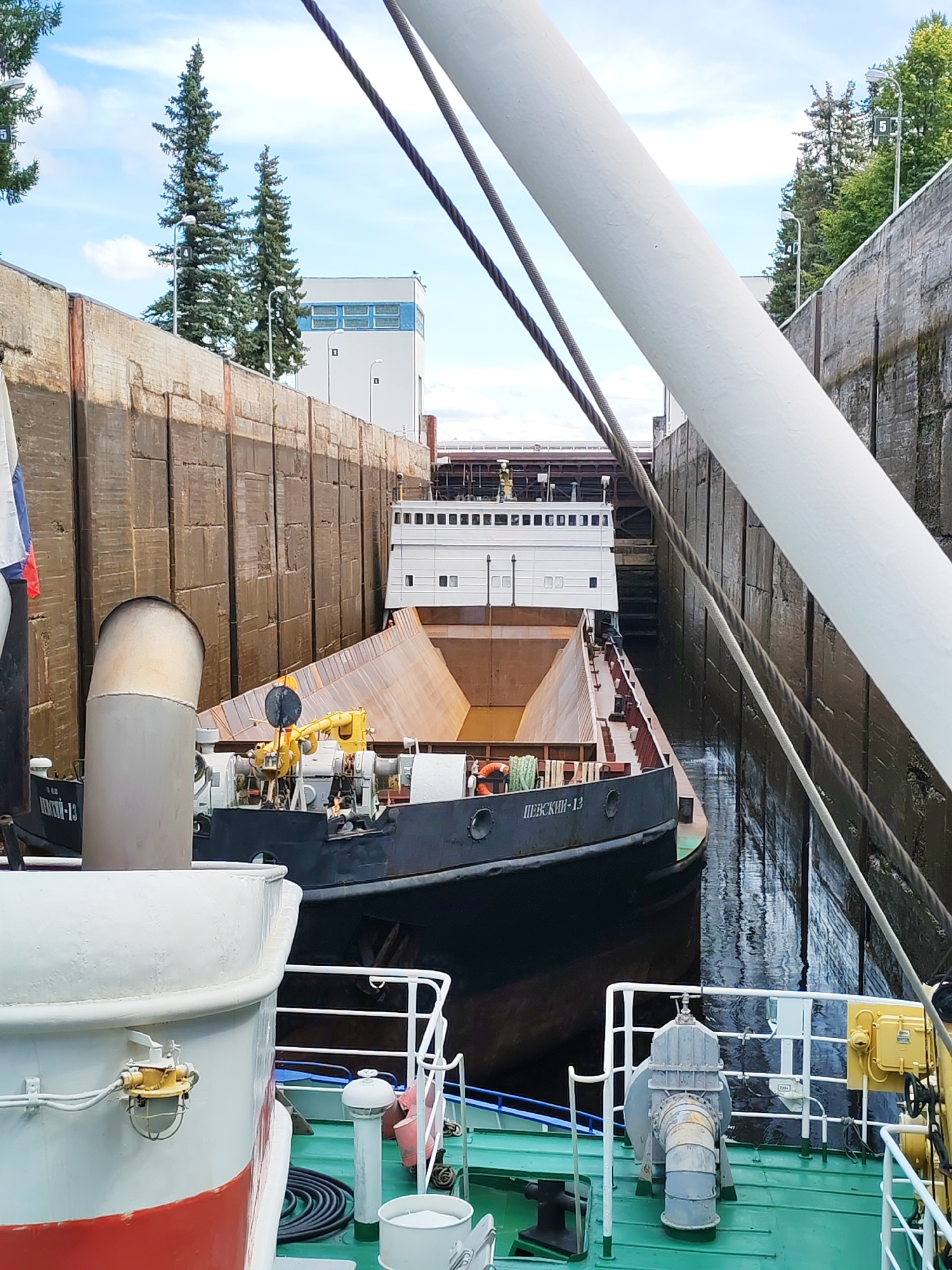 The image depicts a lock chamber in the city of Belousovo, Russia, where two barges are positioned. The foreground shows the deck of a green vessel, partially obscured by a large white cylindrical object. A white railing with vertical supports runs along the edge of the deck. Further into the chamber, a dark-colored barge with Cyrillic lettering reading "ПЕВСКИН-13" is visible, featuring a large, angled, rust-colored cargo hold. Behind this barge, a larger white vessel with multiple windows is docked. The lock chamber walls are constructed from large, textured concrete blocks, with water reflecting the sky and vessels. In the background, trees and a modern building with blue window frames are present under a cloudy sky. The scene suggests the ongoing operation of a lock for maritime transport.