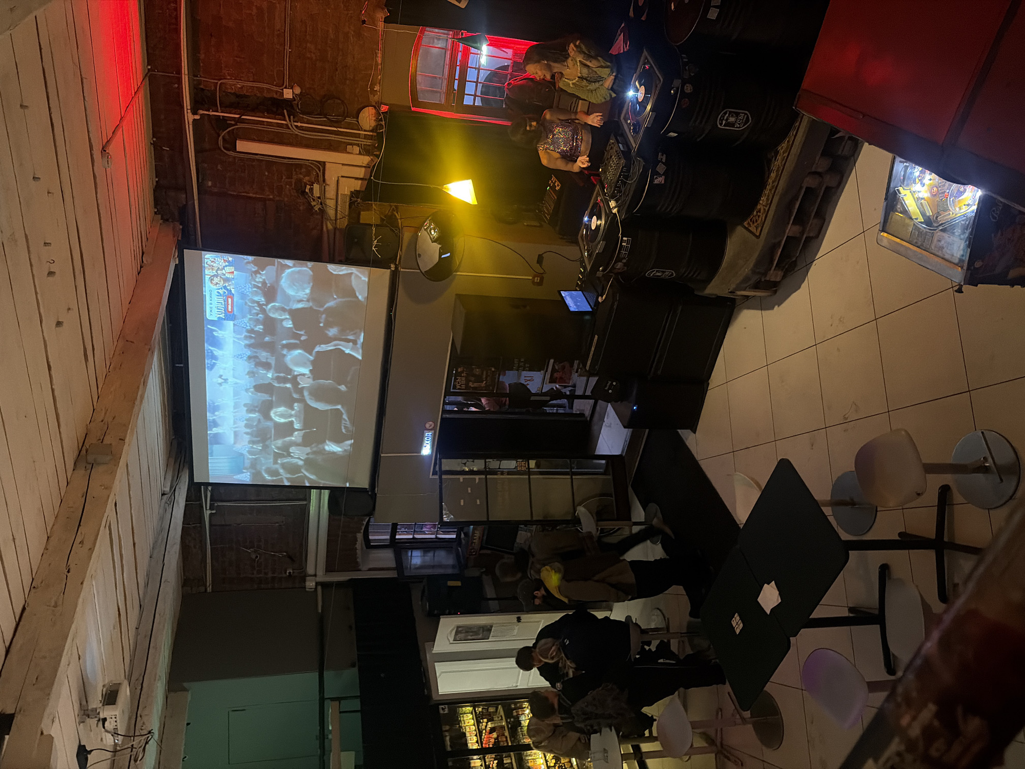 The image depicts an indoor event space, likely a bar or club, in Saint Petersburg, Russia. A large projection screen displays a scene with an audience. In the foreground, a group of people are gathered around a table, with others standing nearby. To the right, two women are positioned behind DJ equipment, suggesting a musical performance is taking place. The environment appears industrial with exposed wooden beams on the ceiling and brick walls. Several barrels are used as bases for the DJ setup and are decorated with stickers. A pinball machine is visible in the lower right corner of the frame. The lighting is dim, with spotlights and red accent lights contributing to the atmosphere.