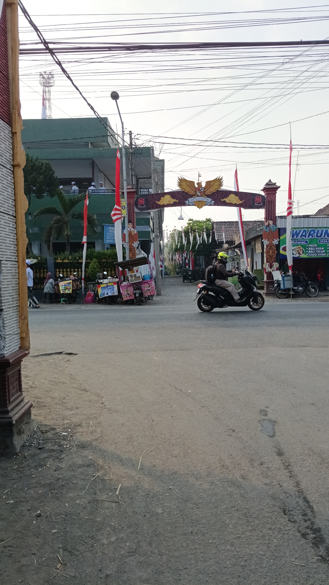 The image captures a street scene in Ploso, Indonesia. An archway, adorned with the Garuda Pancasila symbol and the number "81" on its pillars, spans across the road. Red and white Indonesian flags are prominently displayed on either side of the arch and along the street. A man wearing a yellow helmet and a backpack is riding a black motorcycle from right to left across the frame, passing under the arch. To the left of the arch, a building with green and white signage that includes the word "WARUNG" is visible. In the foreground, a dusty asphalt road occupies the majority of the lower half of the image. Several food carts are parked on the side of the road near the archway, with a few people standing or sitting around them. In the background, more buildings and trees are visible. The sky is overcast with visible power lines crisscrossing the upper portion of the image.