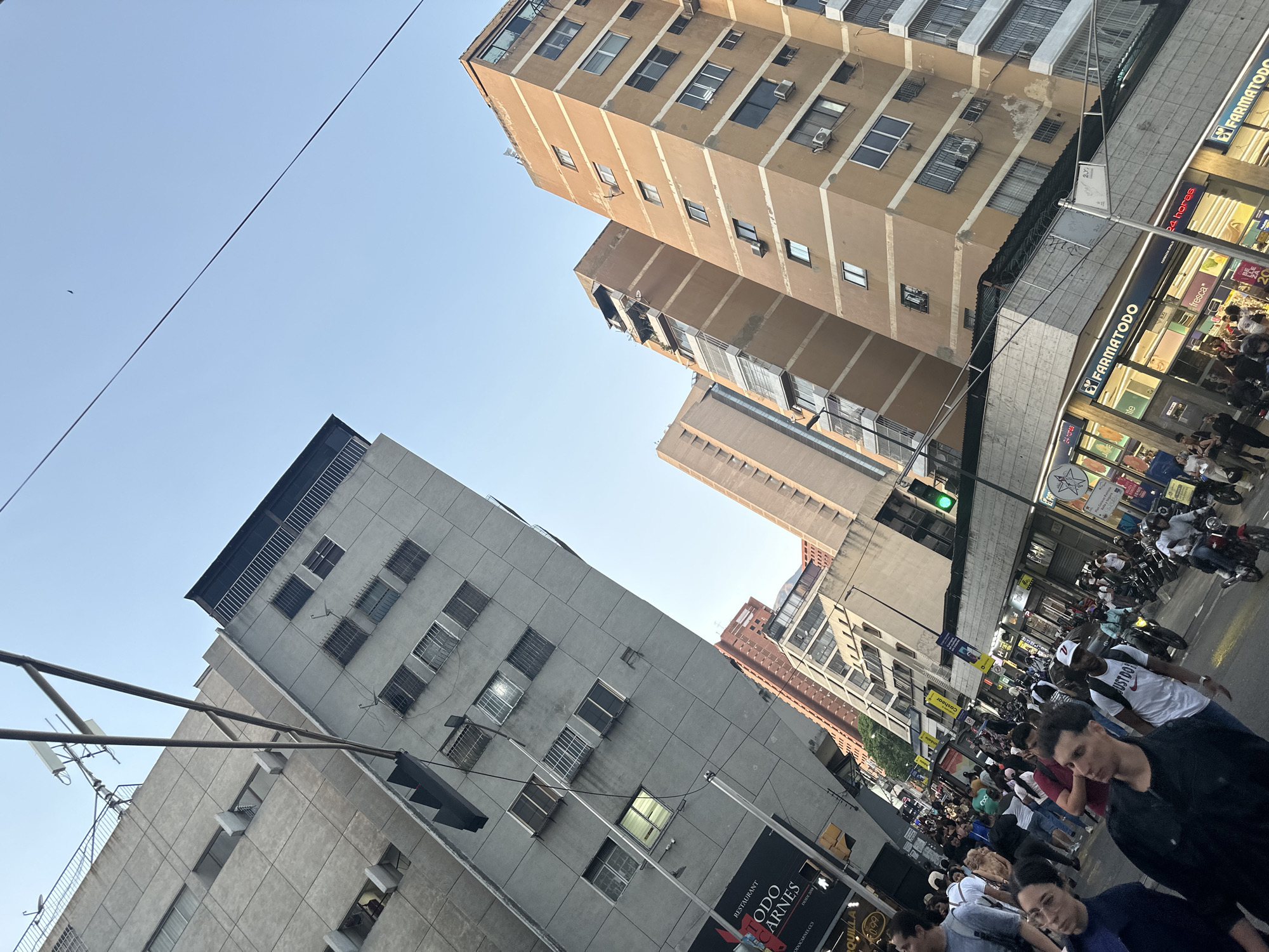 The image captures a street scene in Caracas, Venezuela. Several multi-story buildings are visible, one a beige color with horizontal striping, and another grey. A "Farmatodo" is open for business on the street level. A considerable number of pedestrians are present, some walking and some standing. There are also a number of motorcycle delivery workers with bikes on the sidewalk. Signs for a restaurant called "TODO Carnes" can be seen. Street lights and utility poles are also present. The sky is blue.