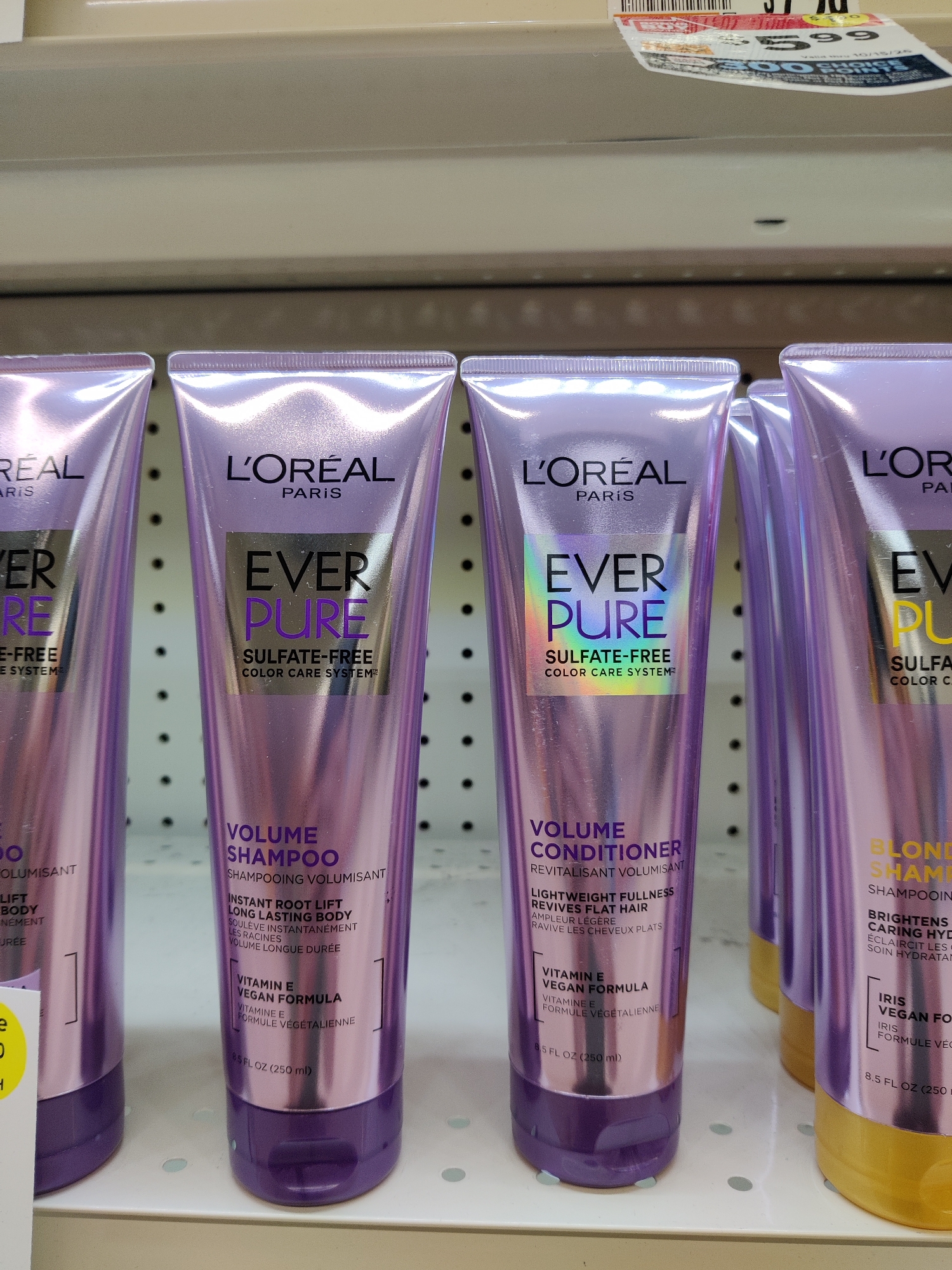 In Mechanicsburg, United States of America, the image depicts a retail store shelf displaying multiple bottles of L'Oréal EverPure hair products. The shelves are white with a beige backing. The bottles are a metallic purple color with white and gold lettering. From left to right, the visible products are: two bottles labeled "L'ORÉAL" with the sub-brand "EVER PURE" and the text "SULFATE-FREE COLOR CARE SYSTEM." The first of these two bottles is partially obscured and has a yellow sticker on it. The next two bottles are also L'Oréal EverPure. One is labeled "VOLUME SHAMPOO" and the other "VOLUME CONDITIONER." Both of these bottles have French translations of their labels. The final visible bottle on the right is mostly obscured, but the L'Oréal EverPure branding is visible along with the text "BLOND SHAMPOO" and "BRIGHTENS CARING HYDRA," also with French translations. The bottles are arranged in a row on a shelf.