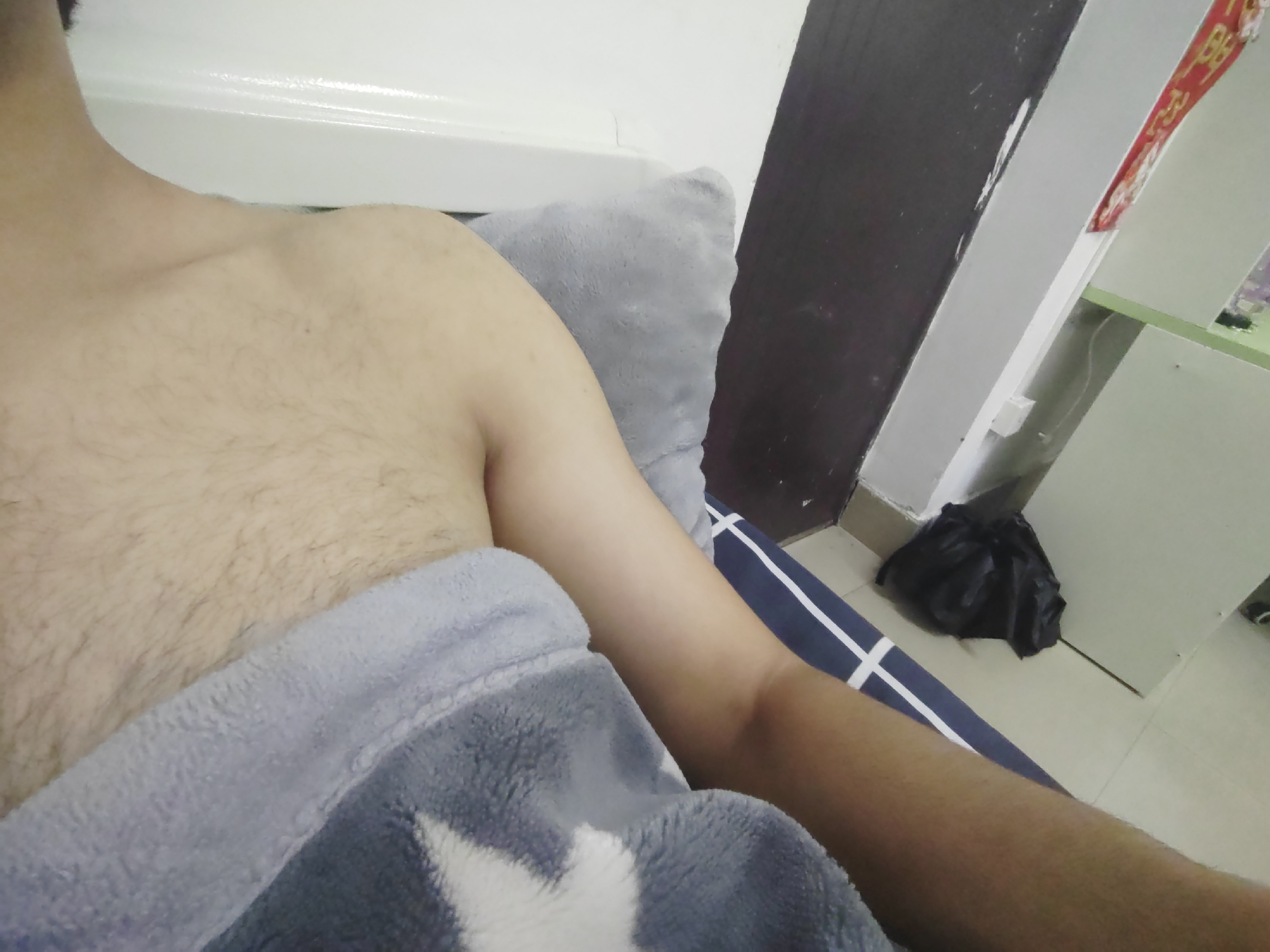 The media file shows a close-up view of a person's torso and arm. The person appears to be lying down, possibly in bed, as indicated by the presence of a pillow and a blanket. The lighting suggests it might be indoors during the day or with artificial light. The setting seems to be a room with a white wall and a dark vertical element, possibly a door or a partition. There is also a black trash bag visible on the floor in the background. A red banner with some text is hanging on the wall in the upper right corner, but the text is not legible. The person's skin is visible, with some body hair on the chest and arm. The overall mood is relaxed and casual, given the comfortable attire and setting.