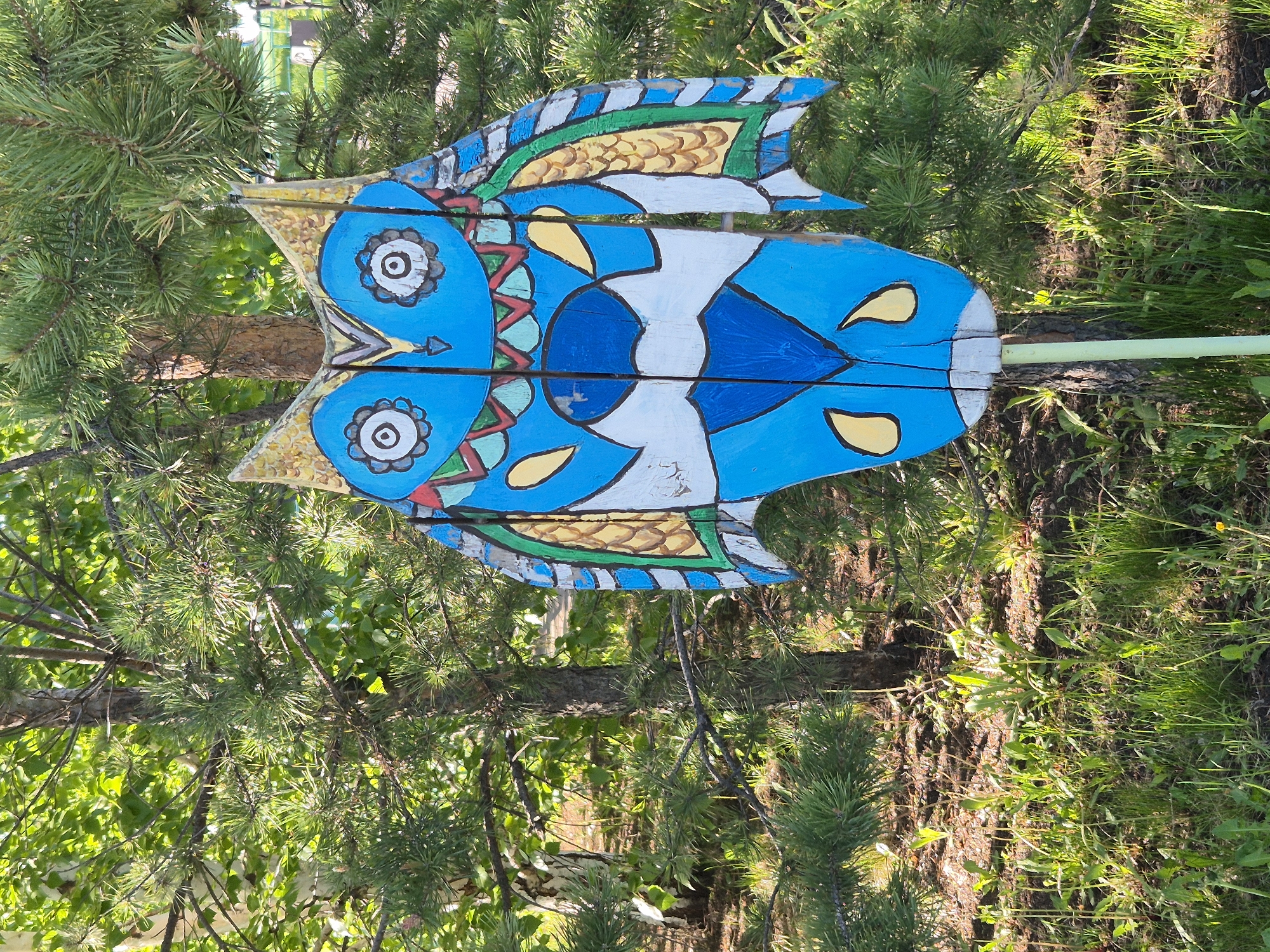 A wooden owl, painted blue with white, yellow, green, and gold details, is mounted on a pole, angled slightly towards the upper left corner of the frame. The owl is positioned amidst a natural setting, featuring green pine branches, grass, and undergrowth. The background suggests a wooded area.
