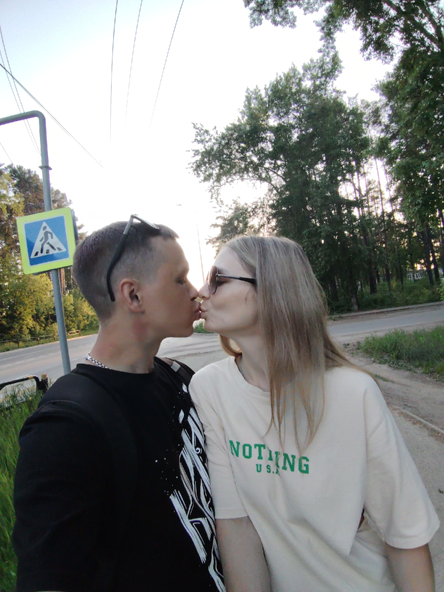 A couple is kissing in front of a pedestrian crossing sign, at an intersection in Chaikovsky, Russia. The man is wearing a black t-shirt with a white design and sunglasses on his head and a silver chain. The woman is wearing a tan t-shirt with the word "NOTHING" printed in green. Sunglasses sit on her face. The photo appears to be taken outdoors, adjacent to a road, with trees visible in the background and a house further in the background. The pedestrian crossing sign is blue and yellow.