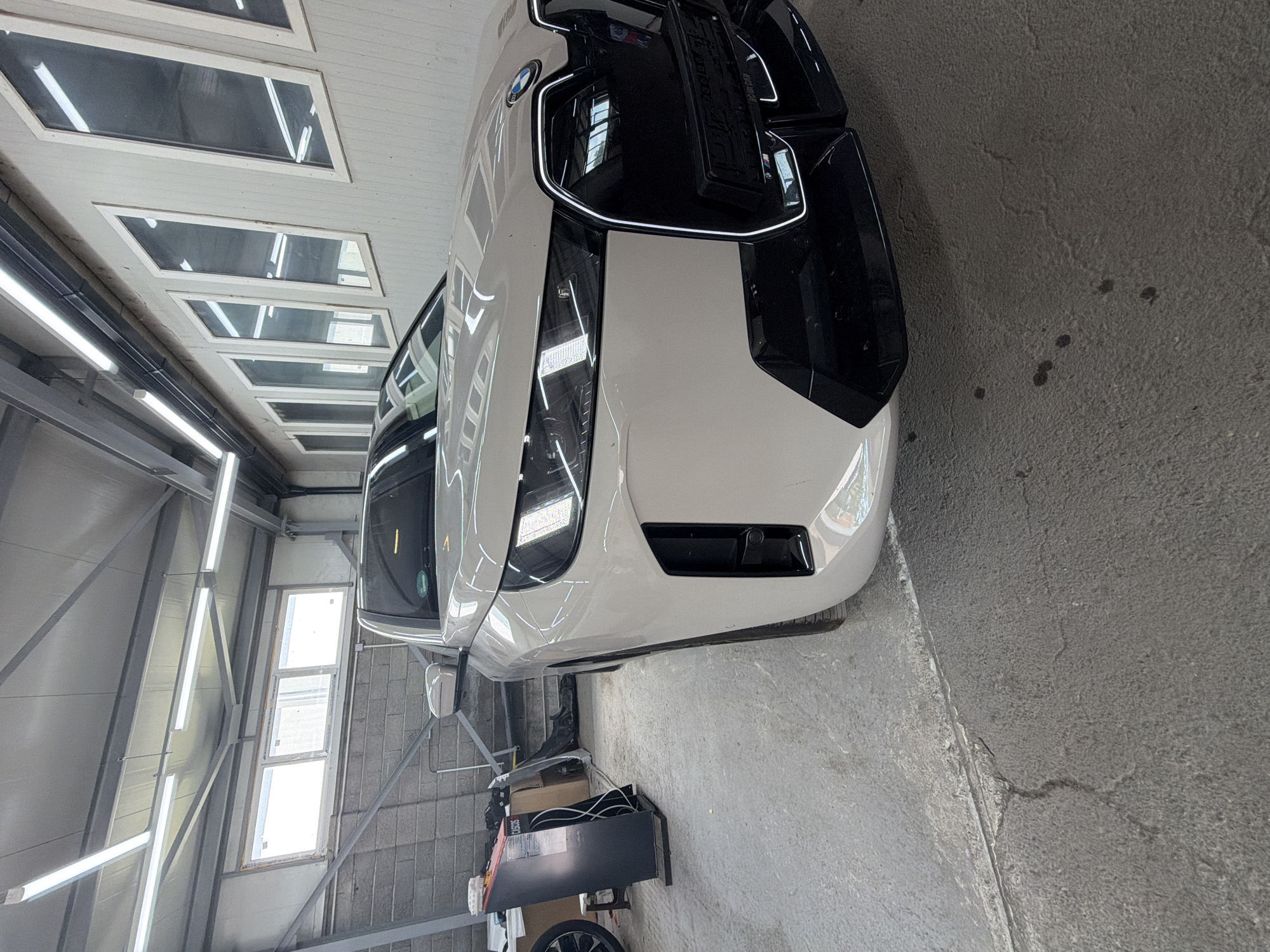 The image shows a white BMW iX parked in a garage or workshop. The car is positioned at an angle, with its front end facing the camera. The garage has a concrete floor with some cracks and stains. Fluorescent lights are mounted on the ceiling, illuminating the space. On the left side of the image, there is a piece of equipment with a screen and some buttons, and to its left, a spare tire is visible. The background shows a brick wall with windows and industrial-style shelving. The image does not contain any people. The location is the city of Burgas, Bulgaria.