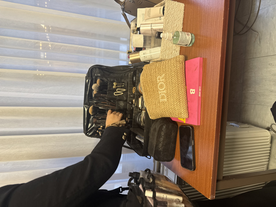 A makeup artist's workstation is captured from above. A black makeup case filled with brushes and cosmetic tools sits open on a wooden surface. To the right are various products, including a woven "DIOR" pouch, a pink "BINNY MATTE" palette, and toiletries in a textured container. A smartphone rests near the edge, while the artist's arm reaches into the makeup case. Suitcase and a black case are seen below on the left.