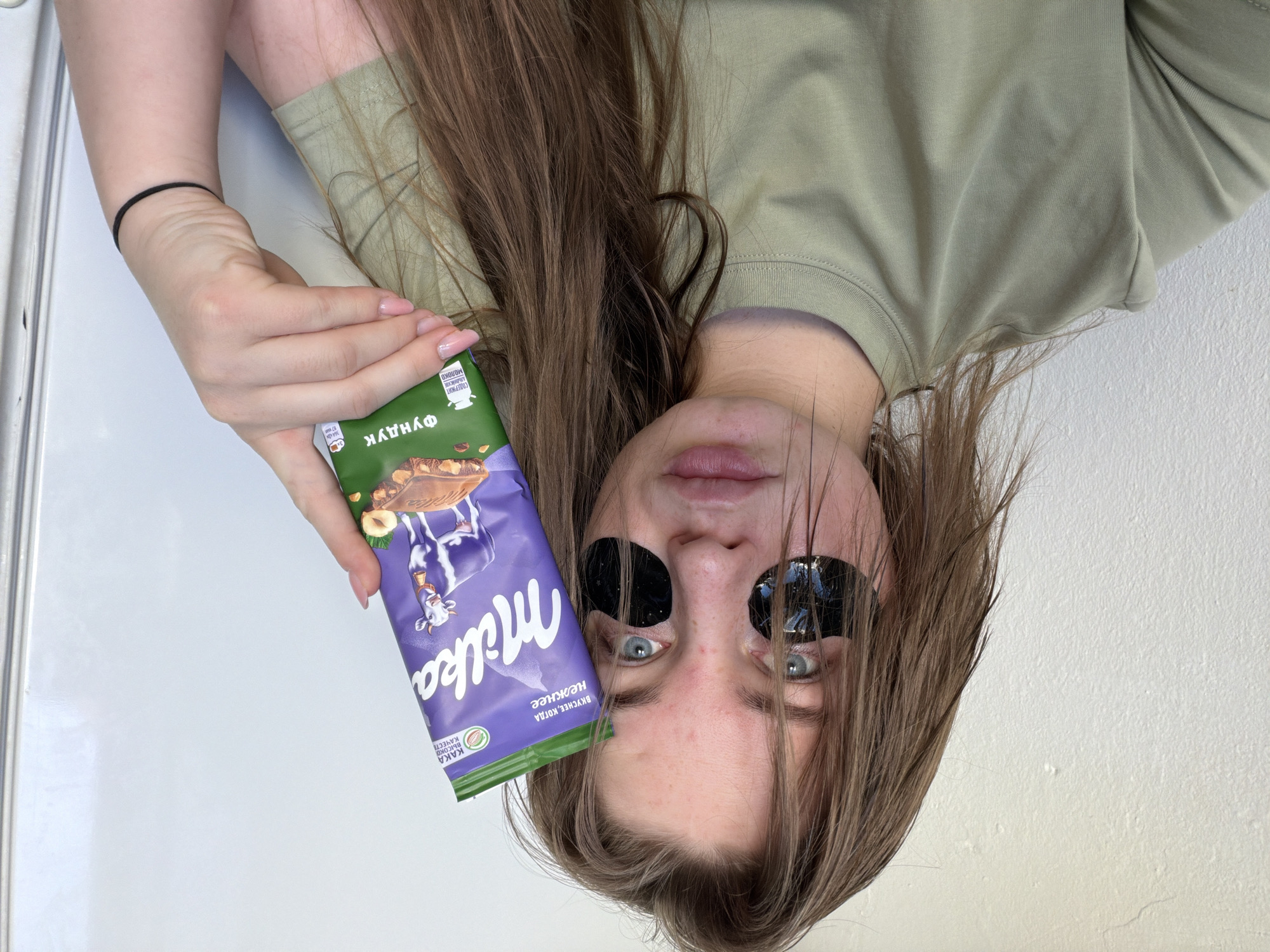 A person, presumably female, is angled at approximately 90 degrees with her head towards the bottom of the image and her torso towards the top. She has light skin, brown hair, and black eye patches. She is wearing a light green, long-sleeved shirt. Her left hand is holding a purple Milka chocolate bar with green accents. The environment appears to be an interior space with white walls and trim. Based on the text on the chocolate wrapper, which includes Russian text, the location is possibly in Russia, potentially in the city of Chaikovsky. There are no other discernible objects or interactions apparent in the cropped scene.