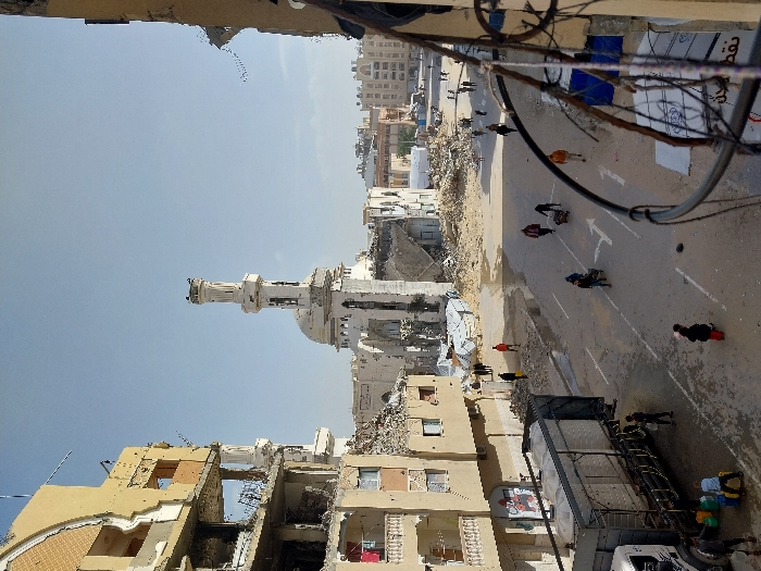 This image depicts a street scene in Khan Yunis, Palestinian Territory, characterized by widespread structural damage. The central subject is a heavily damaged white mosque featuring a tall minaret and a prominent dome, both exhibiting significant structural compromise. To the left, a multi-story building stands partially destroyed, with outer walls collapsed, exposing internal rooms and debris. Another damaged building is partially visible on the right foreground, framed by a metal railing.
The paved street, marked with white directional arrows and lines, shows patches of wetness. Numerous individuals are present on the street and sidewalks. On the left side of the street, a white truck equipped with two large white water tanks is parked, serving as a water distribution point. Several people, including children, are gathered around the truck, actively filling yellow and green jerry cans from spigots. Further along the street, other individuals walk, and several are seen pushing handcarts or small trolleys, possibly transporting goods or supplies. Piles of rubble and debris are scattered across the scene, particularly in areas where buildings have collapsed, and makeshift shelters constructed from tarpaulins are visible in the background among the ruins. The sky is clear, indicating daylight.