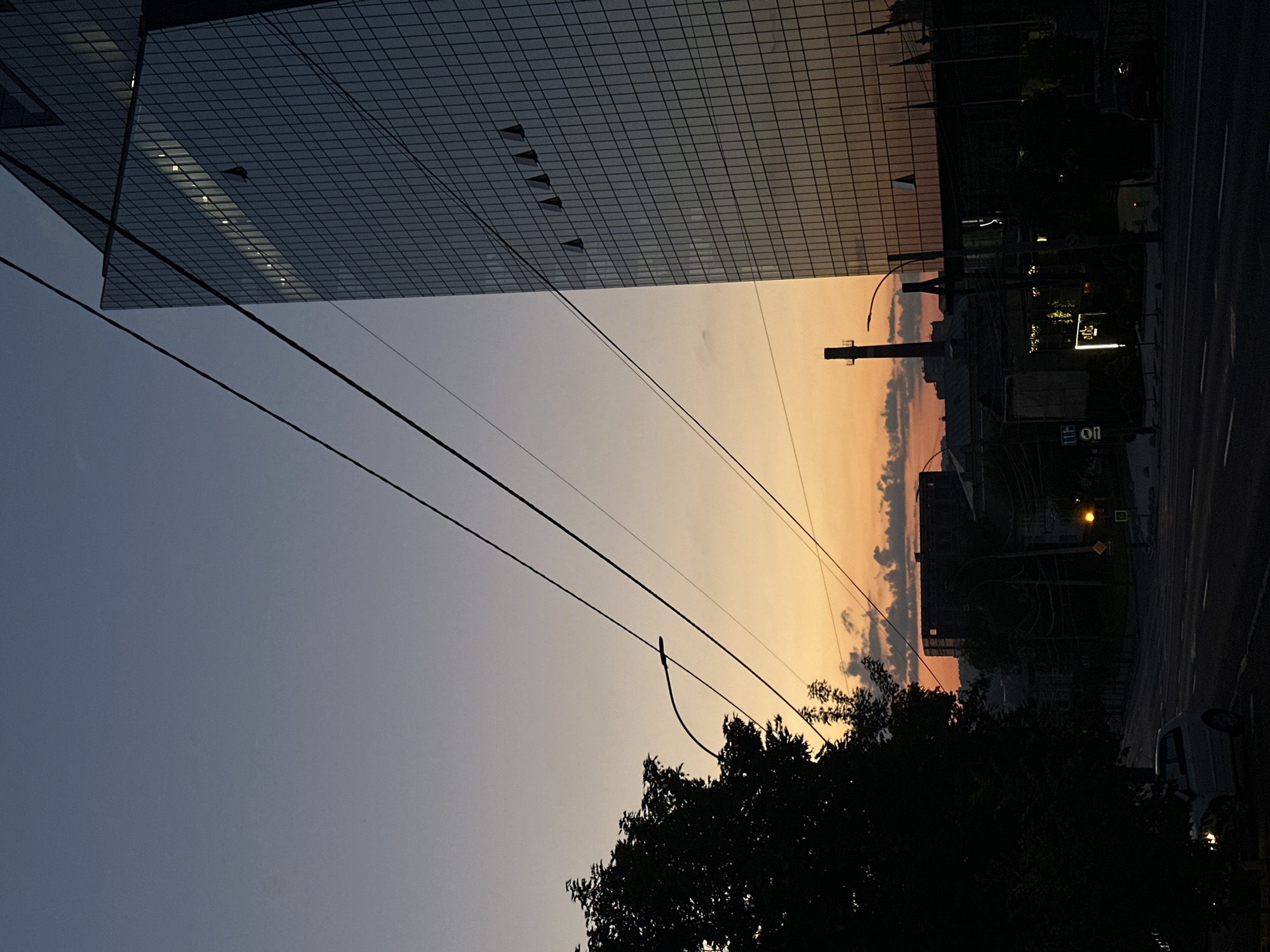 The image captures a city skyline at dusk, likely in Chișinău, Moldova, given the architecture. A tall, modern glass-facaded skyscraper dominates the right side of the frame, its reflective surface mirroring the warm tones of the sunset. Several power lines crisscross the sky in the foreground, creating a geometric pattern against the gradient of the twilight sky, which transitions from a deep blue at the top to shades of orange and yellow near the horizon.
To the left, a silhouetted deciduous tree provides a stark contrast to the illuminated building, its dark foliage obscuring a portion of the sky. In the mid-ground, various low-rise buildings and industrial structures are visible, including a prominent chimney stack. Streetlights and building lights begin to illuminate the scene, adding points of artificial light. A street stretches into the distance, with a white car parked on the side and traffic markings visible on the asphalt. The overall atmosphere is one of urban transition from day to night, with the natural beauty of the sunset juxtaposed against the man-made structures of the city.