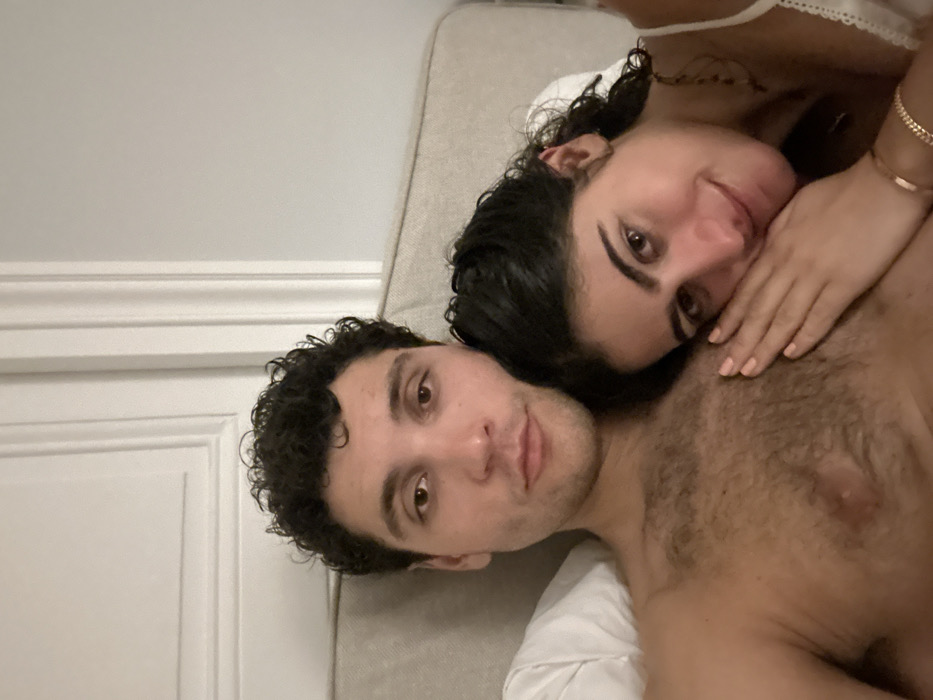 A man and a woman are laying down in bed. The woman rests her head on the man's chest, with her hand gently placed on his skin. Both look at the camera. The man has dark, curly hair and a fair complexion. The woman has dark hair, bracelets, and a tattoo on her shoulder. They are on a light-colored mattress with a white sheet visible. A white wall and door trim are visible on the left side of the frame.