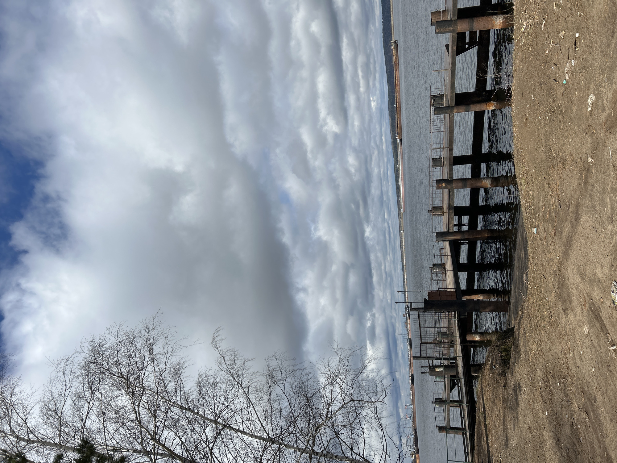 The image depicts a waterfront scene in Chaikovsky, Russia. The sky is filled with dense, striated clouds, with a small patch of blue visible in the upper left corner. A bare tree with thin branches occupies the lower left of the frame. To the right, a dark body of water, presumably the Kama Reservoir, is partially obscured by a dilapidated pier structure consisting of rusted metal pilings and sections of decaying wood. The shoreline is composed of rough, uneven dirt and debris. In the distance, the skyline of Chaikovsky is faintly visible. No people are present in the image.