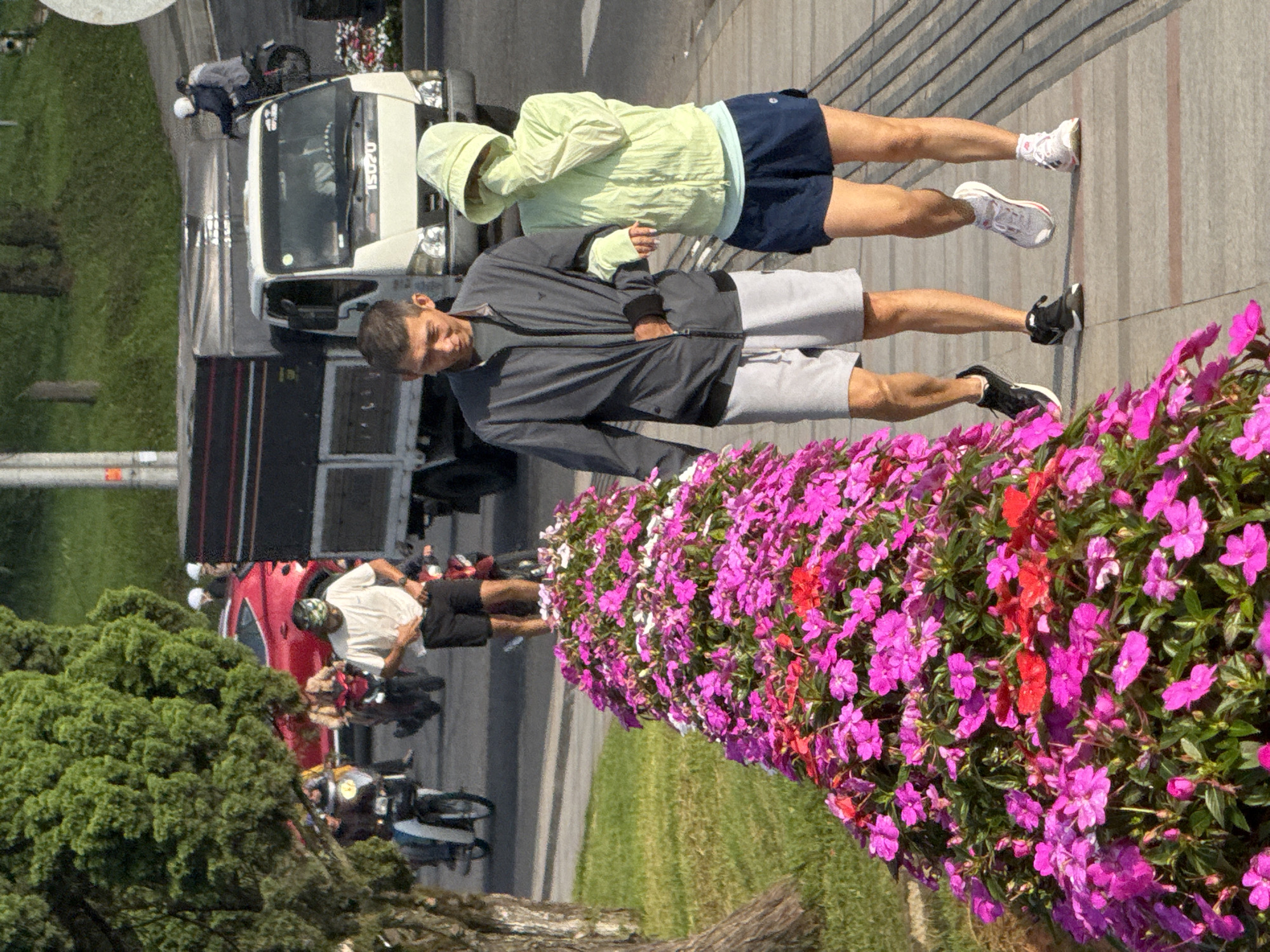 This image captures a street scene in Da Lat, Vietnam, featuring a couple walking along a sidewalk lined with a dense bed of vibrant pink and red impatiens flowers. The man on the left, wearing a grey jacket and grey shorts, has his arm around the woman. The woman on the right, dressed in a light green hooded jacket and dark blue shorts, shields her face with her jacket sleeve. Behind them, a white Isuzu truck is parked, and to the left, a portion of a red car and a lush green tree are visible. In the background, several people on motorcycles and a truck are present, indicating traffic activity. The ground is paved with a light-colored sidewalk, and a grassy area is adjacent to the flower bed.