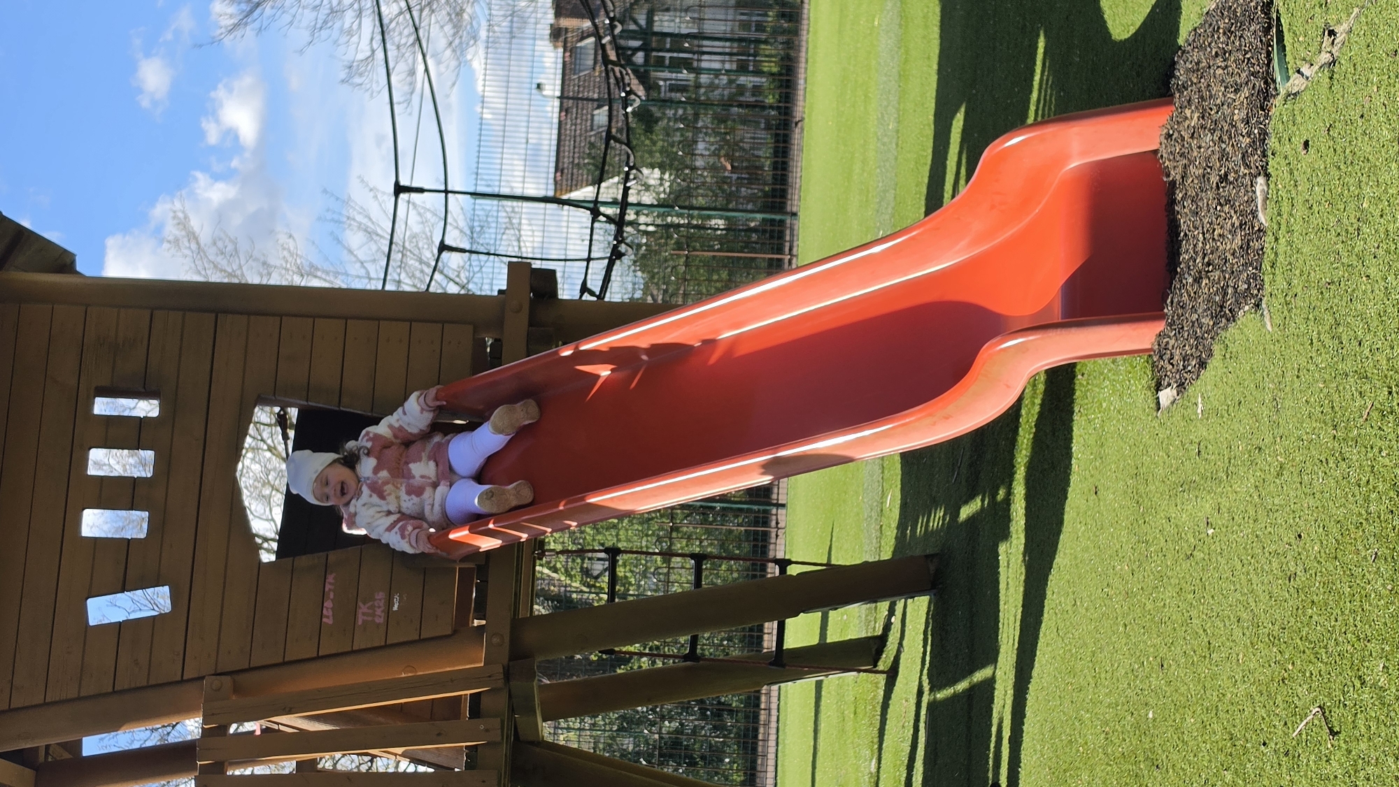 A young child in a patterned jacket and white leggings and hat is sliding down a red slide that is attached to a wooden play structure. The slide exits onto a green surface with what appears to be shredded rubber or bark mulch adjacent to the slide exit. In the background there are buildings, fences, and trees. The photograph was taken outdoors on a sunny day. The specific location within London is not apparent from the image.
