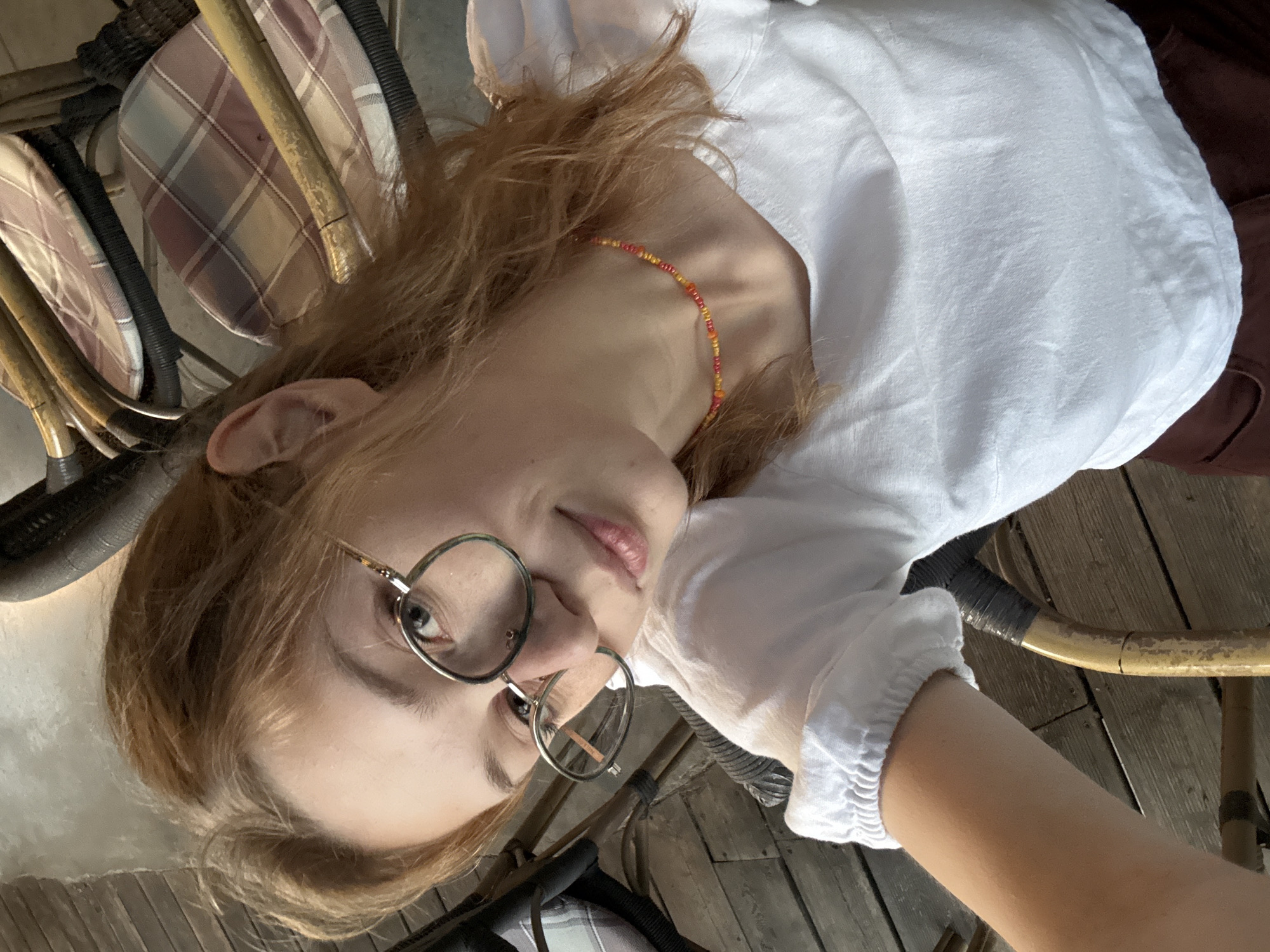 A young person with light brown hair and glasses is looking directly at the camera. They are wearing a white shirt and a beaded necklace of orange and yellow beads. The person is sitting in a chair. The chair has a patterned cushion. The floor is wooden. The location is not identified as Kragujevac, Serbia.