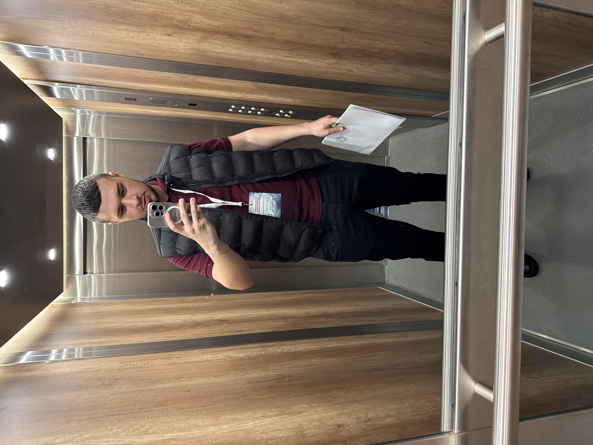 A person stands inside an elevator, taking a selfie with a smartphone. He is wearing a maroon shirt, a black vest, and black pants. He holds a piece of white paper in his left hand. The elevator's interior consists of wood-panelled walls and a control panel with buttons. The floor appears to be grey. The person is also wearing a lanyard with a tag, although the text on the tag is not clear. The elevator has a metal handrail on one side. Without additional information, the location in Sofia, Bulgaria cannot be determined.
