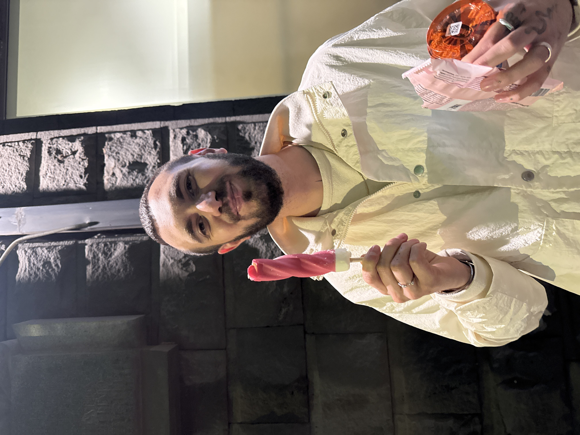 A man stands outdoors, holding a pink and white swirled popsicle in his left hand. In his right hand, he holds an open package and a small orange container. He wears a light-colored jacket over a cream-colored sweater. He has dark hair and a beard. The background includes a dark gray stone or brick wall with what appears to be a window above. The setting is likely urban. This picture was taken in Yerevan, Armenia.