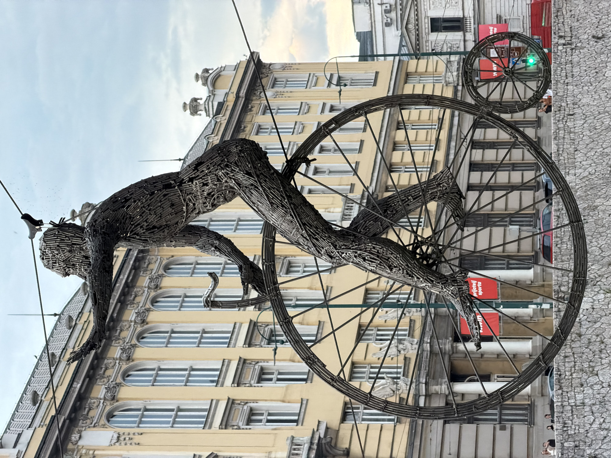A metal sculpture of a person riding a vintage bicycle is prominently displayed in Sarajevo, Bosnia and Herzegovina. The sculpture, made of intricately woven metal rods, depicts a figure with outstretched arms, poised dynamically on a large front wheel of a penny-farthing style bicycle. The bicycle's frame and wheels are also constructed from metal rods, creating a skeletal and artistic effect.
The sculpture is positioned in front of a grand, light yellow building with multiple arched windows and classical architectural details, suggesting a historical or institutional setting. The building has a mansard roof with dormer windows. In the background, other buildings are visible, including one with a white facade and classical columns.
Below the sculpture, a stone wall, possibly a bridge parapet or embankment, lines the foreground. A street runs behind this wall, where a few vehicles, including a red car and a red SUV, are present. Several people are casually walking or standing near the wall and on the street, their presence providing a sense of scale to the artwork.
The sky above is overcast with diffused light, indicating it is likely daytime, possibly late afternoon or early morning. Overhead wires and a lamppost are also visible, suggesting an urban environment. Two red banners with text are hung on the yellow building, though the text is not fully legible. A traffic light with a green signal is visible to the right of the sculpture's smaller rear wheel. The sculpture is a striking public art installation, adding a unique artistic element to the cityscape of Sarajevo.