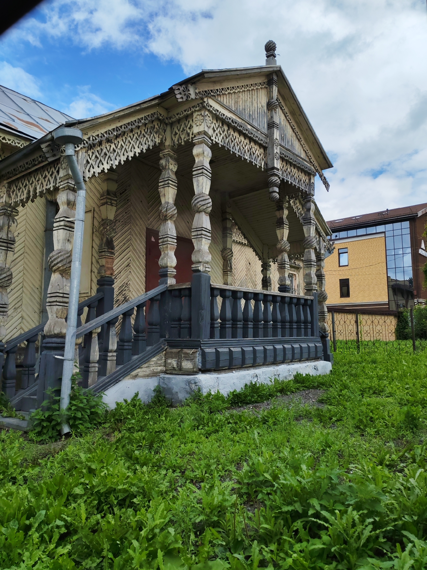 The image depicts a weathered, two-story wooden house with intricate carvings on its porch. The house is painted a light yellow and shows signs of age, with peeling paint and exposed wood. A dark gray railing with balusters lines the porch. A metal downspout runs down the side of the house. In the foreground, lush green grass and foliage grow. Behind the wooden house, a modern building with yellow brick and large glass windows is visible, separated by a black metal fence. The sky above is partly cloudy with patches of blue. The scene is set in Bol'shie Mkhi, Russia.