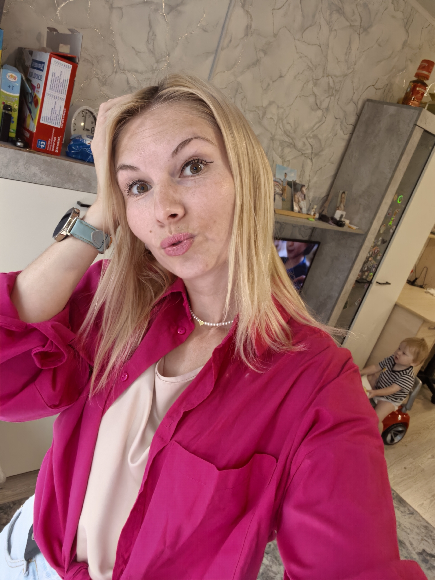 A woman with blonde hair and fair skin, wearing a bright pink shirt over a beige top and a pearl necklace with a heart pendant, takes a selfie. She has her left hand on her head and is looking at the camera with her lips pursed. A smartwatch with a light blue band is visible on her left wrist. In the background, a toddler wearing a striped shirt and a diaper is sitting on a red ride-on toy. The environment appears to be an indoor space with shelves displaying various items, including boxes and framed pictures. The wall has a marble-like texture. The scene does not provide any specific landmarks or indicators to suggest the location is Sterlitamak, Russia.