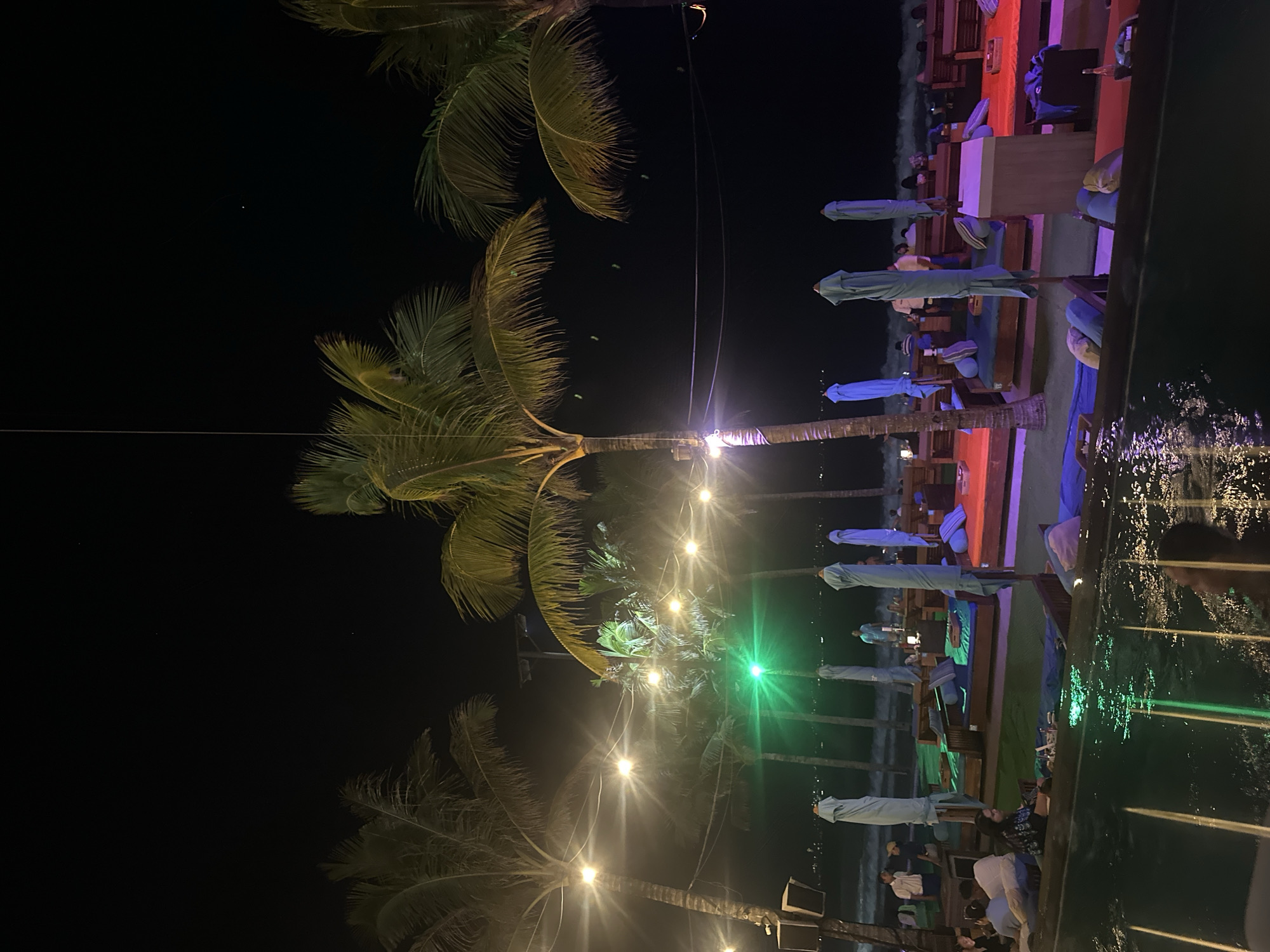 This media file captures a nighttime scene of an outdoor restaurant or bar situated along a body of water, likely the ocean, in North Kuta, Indonesia. The image is dominated by several tall palm trees, their fronds visible against the dark sky, especially on the left side of the frame.
A string of warm, glowing Edison-style lights stretches horizontally across the middle of the image, strung between the palm trees, providing the primary illumination. A distinct green light source is also visible further down, casting a bright green reflection onto the water.
Below the palm trees and lights, a series of outdoor seating arrangements are visible, set on what appears to be a wooden deck or patio next to the water. These include wooden tables and padded loungers, many featuring light blue cushions and closed light blue umbrellas. The seating areas are illuminated with various colored ambient lights, predominantly purple, pink, and green, creating a relaxed atmosphere.
Several people are seated at these tables and loungers, engaged in what appears to be dining, socializing, or simply relaxing. Their forms are somewhat indistinct due to the low light.
To the right, a dark body of water shows ripples and reflections of the artificial lights from the establishment and possibly distant sources. The sky above is dark and clear, indicating a pleasant evening. There is no visible text in the image. The overall setting suggests a tranquil and inviting tropical resort or beach club environment.