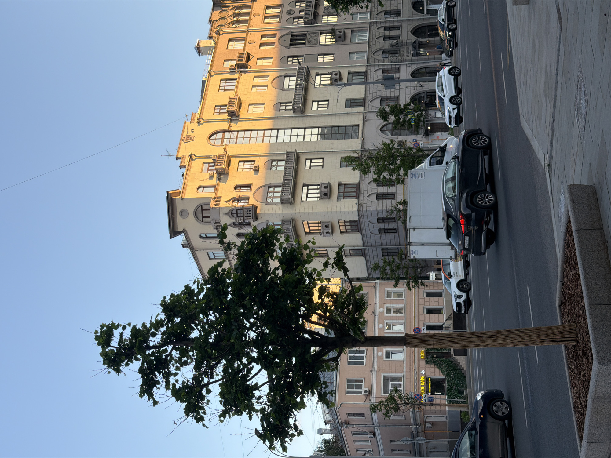 A daytime scene on a Moscow street presents a multi-story building with cream-colored facade and architectural details on the left side of the frame, partly obscured by a large tree. The street is active with vehicular traffic, including several cars and a white box truck. The vehicles are aligned along the road's edge, suggesting parked or slow-moving conditions. A sidewalk is visible to the right, demarcated by a curb and an adjacent strip of landscaping. The lighting is bright, casting shadows that indicate the sun's position. Signs in Russian are also present, reinforcing the location as Moscow.
