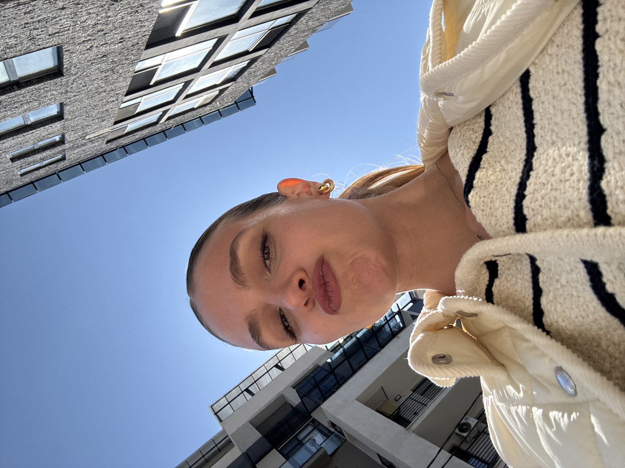 A person is looking down at the camera, with buildings in the background under a blue sky. The buildings appear modern with glass windows and balconies. The person has fair skin, brown eyes, and dark lipstick. They are wearing a jacket with vertical black stripes. A gold earring is visible in the person's ear. The location is likely Tbilisi, Georgia, based on the architectural style of the buildings.