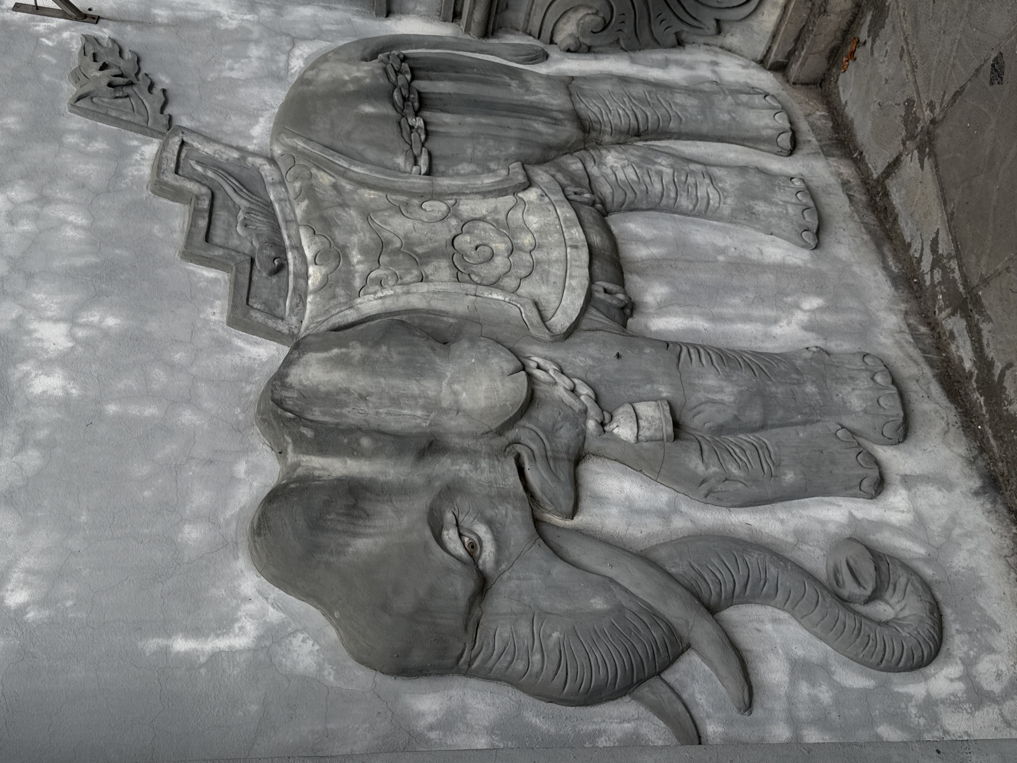 The image shows a gray, intricately carved stone relief of an elephant lying on its side. The elephant is adorned with a decorative harness and a chain. The relief appears to be part of a building facade or wall in Hà Nội, Vietnam, judging by the architectural style. The background is a textured, gray surface, possibly concrete.