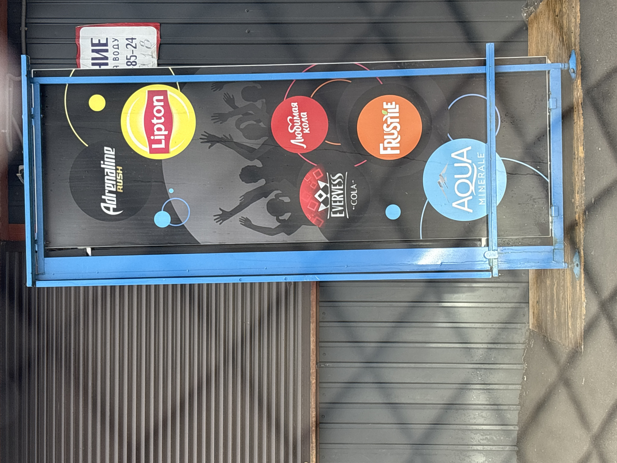 Here's a description of the image:
The image depicts an advertisement sign in Ust-Ordynsky, Russia. The sign has a blue frame. The background is dark, with silhouetted figures. There are brand logos, including "Adrenaline Rush," "Lipton," "EVERVESS - COLA," "Любимая кола" (Beloved Cola), "AQUA MINERALE," and "FRUSTILE." Above the sign, there is a partial sign in Russian, possibly indicating "НИЕ," "Я ВОДУ" and "85-24". The sign is mounted on a wall with a dark metal roll-up door and a gray horizontal panel wall. The adjacent ground area shows traces of moisture.