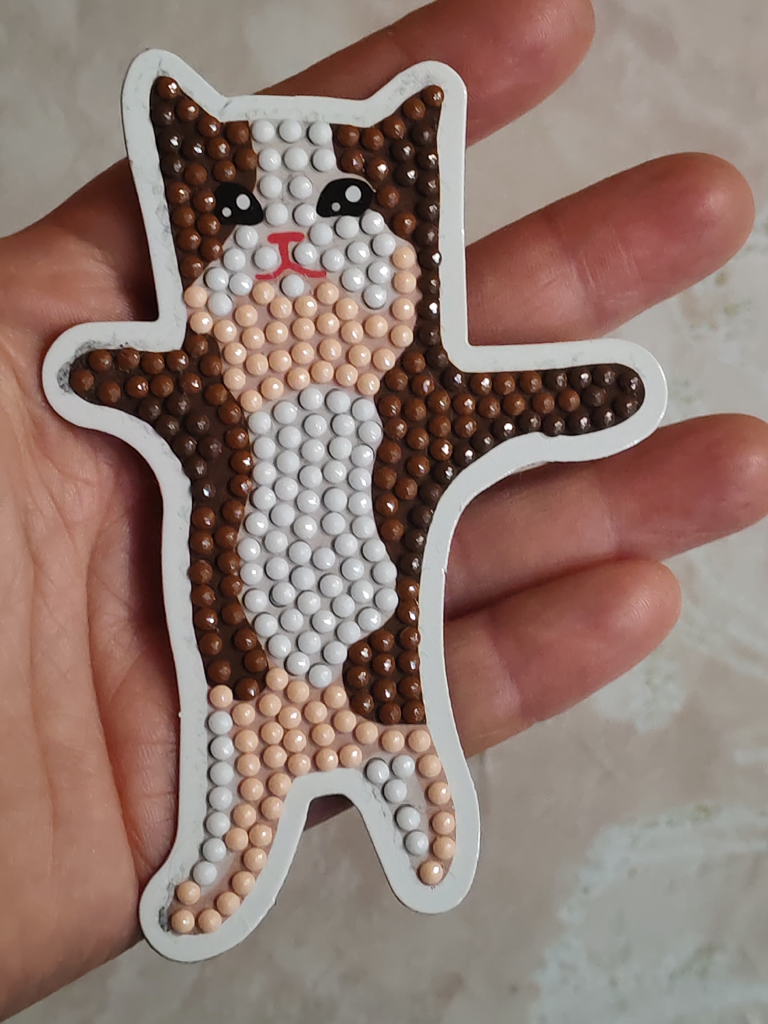 A hand holds a diamond art sticker depicting a cartoon cat. The cat has a white body with brown and tan patches. It has a red nose and black eyes. The sticker has a white border. The background is a blurry mix of skin tones and a cream-colored surface.