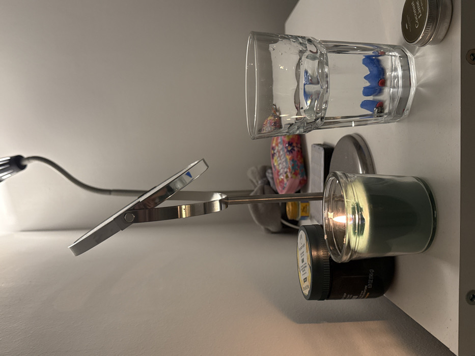 A clear glass of water sits on a white shelf, its contents partially obscured by colorful objects. Below it, a candle in a glass jar is lit, casting a warm glow. A black container sits beneath the candle. A silver clip lamp extends towards the left, its head tilted upwards. The scene is softly lit, highlighting the various textures and shapes of the objects. The background is a plain white wall.