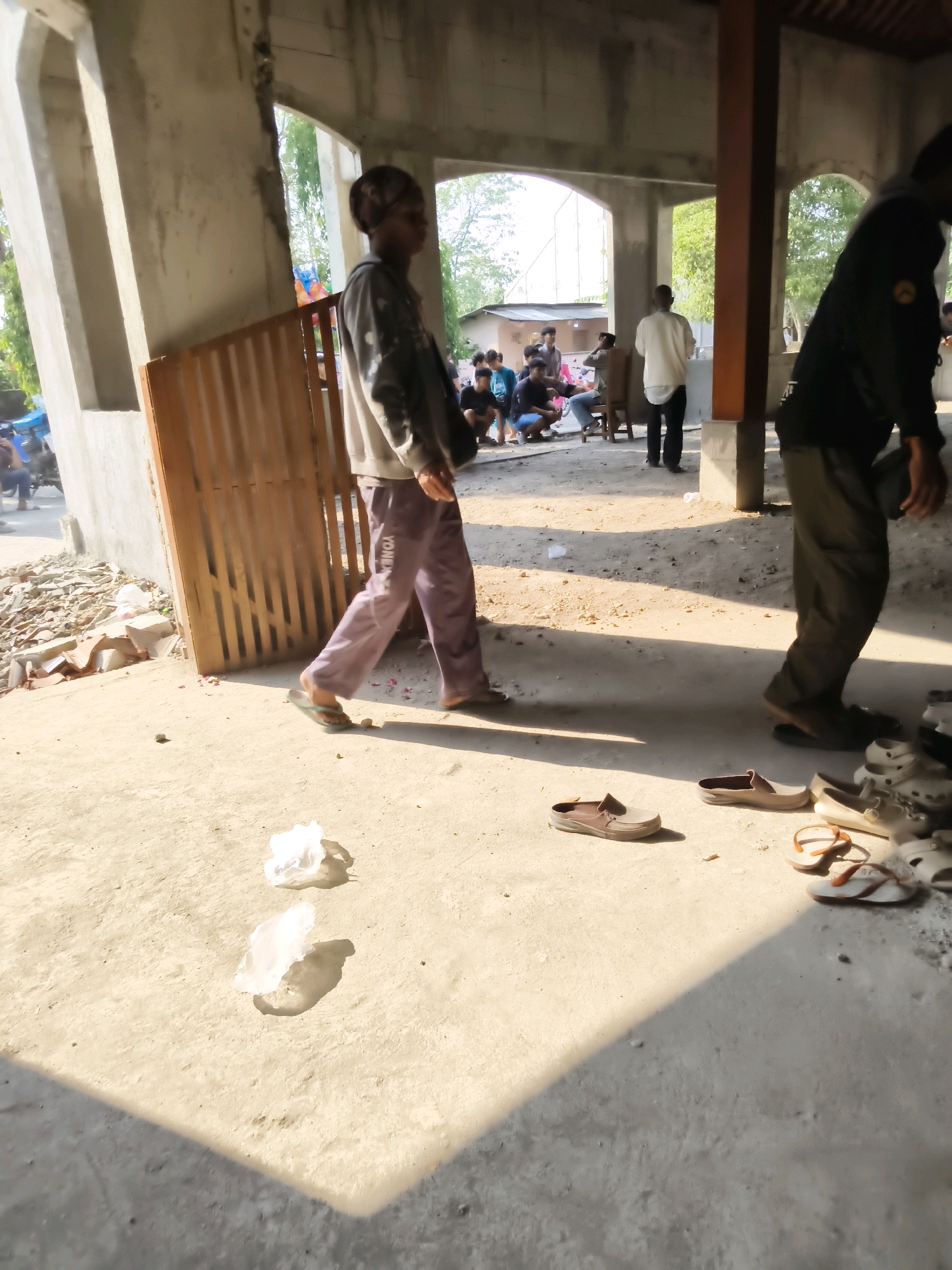 In Kedungan, Indonesia, a person in purple pants and a grey hoodie walks from right to left in the foreground, casting a shadow on the concrete floor. To the left, a wooden gate stands near concrete pillars. In the background, several individuals are gathered, some seated and others standing. A pile of construction debris is visible on the far left. Various footwear, including sandals and closed-toe shoes, are scattered on the ground. The scene appears to be outdoors, possibly near a structure under construction or renovation.