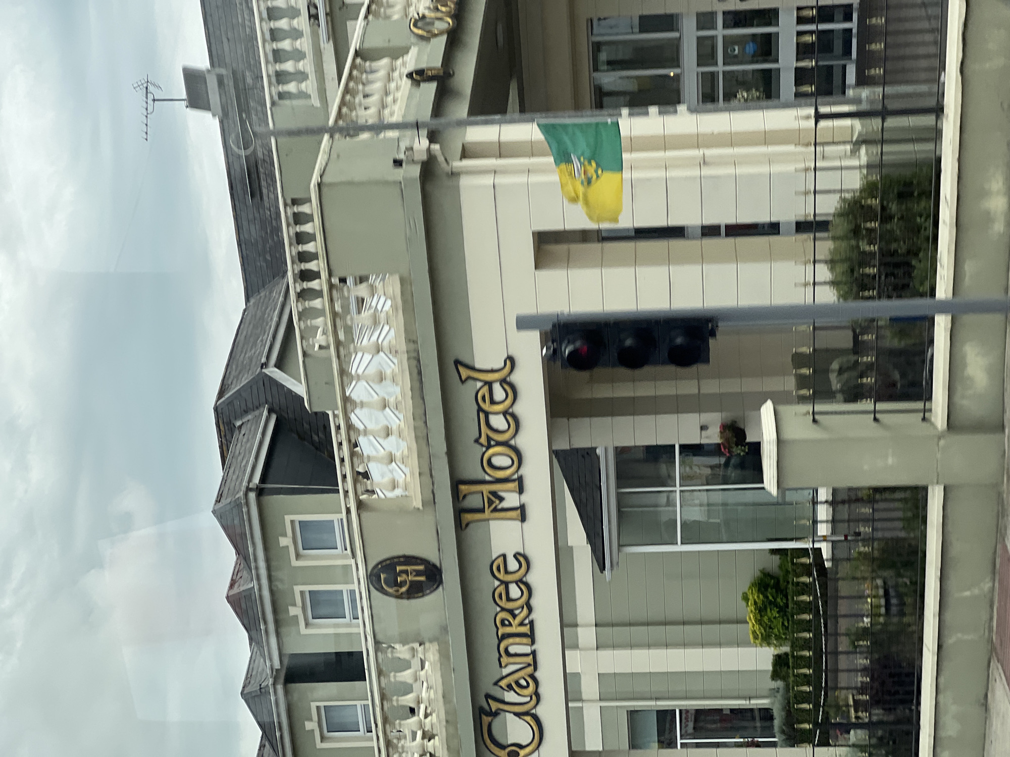 This image depicts the Clanree Hotel in Letterkenny, Ireland. The hotel is a light green building with a white trim, featuring multiple windows and a sloped, dark grey roof. Above the entrance, a prominent gold sign reads "Clanree Hotel". A circular emblem with the initials "CH" is visible on the upper facade.
In front of the hotel, a black metal fence encloses a landscaped area with hedges and small plants. A traffic light with red and black signals stands to the left of the entrance. A flag, split horizontally between yellow and green with a crest in the middle, waves from a flagpole attached to the building. The sky is overcast with grey clouds.