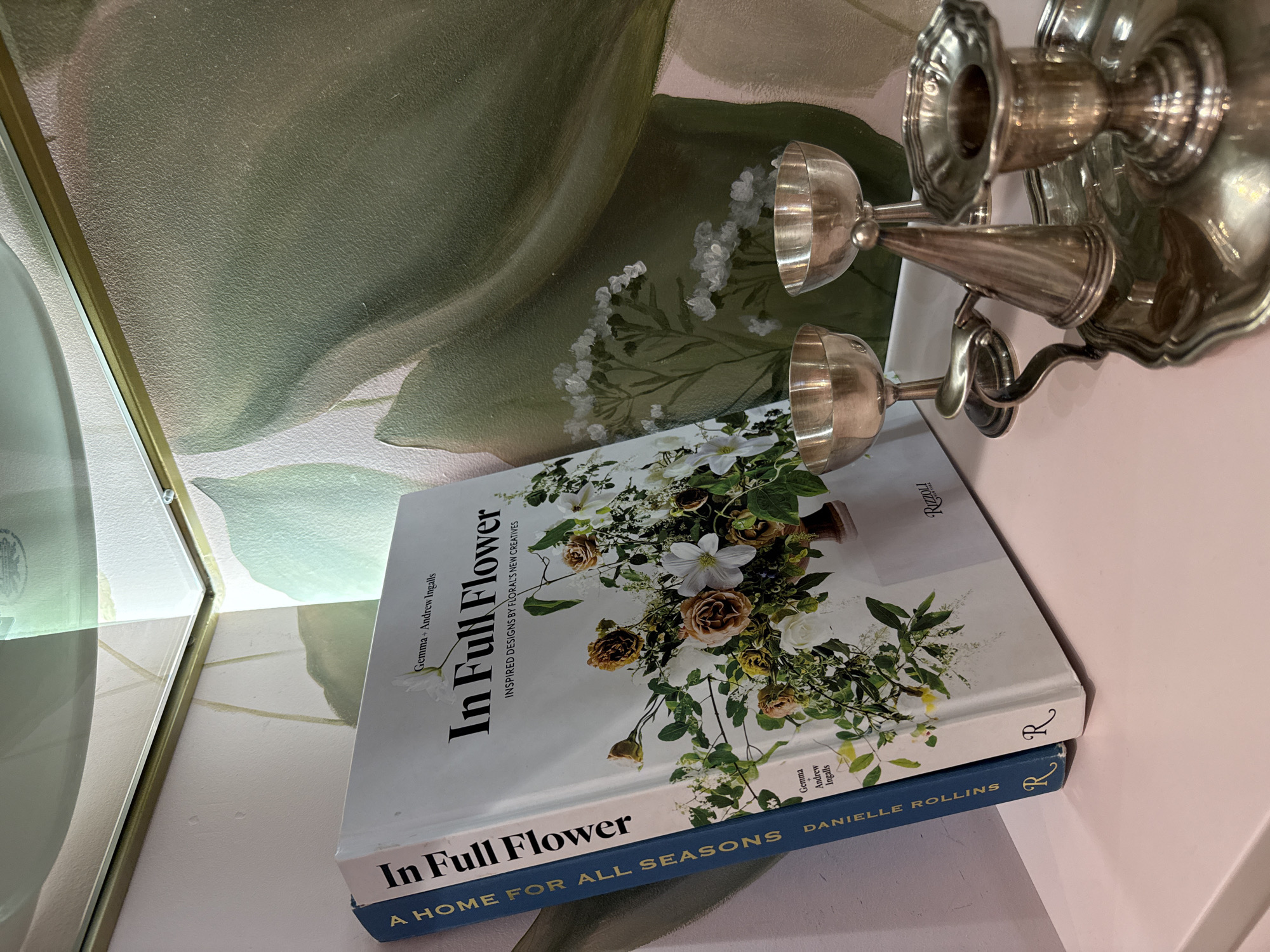 The image shows two books standing upright on a pale pink surface. The book on the right has a white cover with the title "In Full Flower" prominently displayed in black text. Below the title, it reads "INSPIRED DESIGNS BY FLORAL'S NEW CREATIVES." The cover features a floral arrangement with various flowers and greenery. The names "Gemma + Andrew Ingalls" are visible above the title, and "Gemma Andrew Ingalls" is repeated in smaller text on the lower right corner. The spine of this book is white with a black "R" logo.
The book on the left has a dark blue spine with white text that reads "A HOME FOR ALL SEASONS" stacked vertically, followed by "DANIELLE ROLLINS" in smaller text. A stylized black "R" logo is also present on the spine.
In front of the books, on the same surface, are several silver objects. Two small silver chalice-like cups are positioned to the right and center. To the right of these cups is a silver tray with a decorative edge. On the tray, there is a candlestick holder with a central stem and a flared base. A conical silver object, possibly a snuffer or a decorative finial, is placed in front of the candlestick.
The background consists of a wall with a light-colored textured surface and a large, abstract mural of green leaves. The corner where two walls meet is visible in the upper left of the image, with a golden frame and a reflective surface above it. The lighting suggests an indoor setting, possibly a shelf or a tabletop in a room decorated with the floral mural.
There is no discernible information within the image that would suggest a specific location in the city of Kyiv, Ukraine.