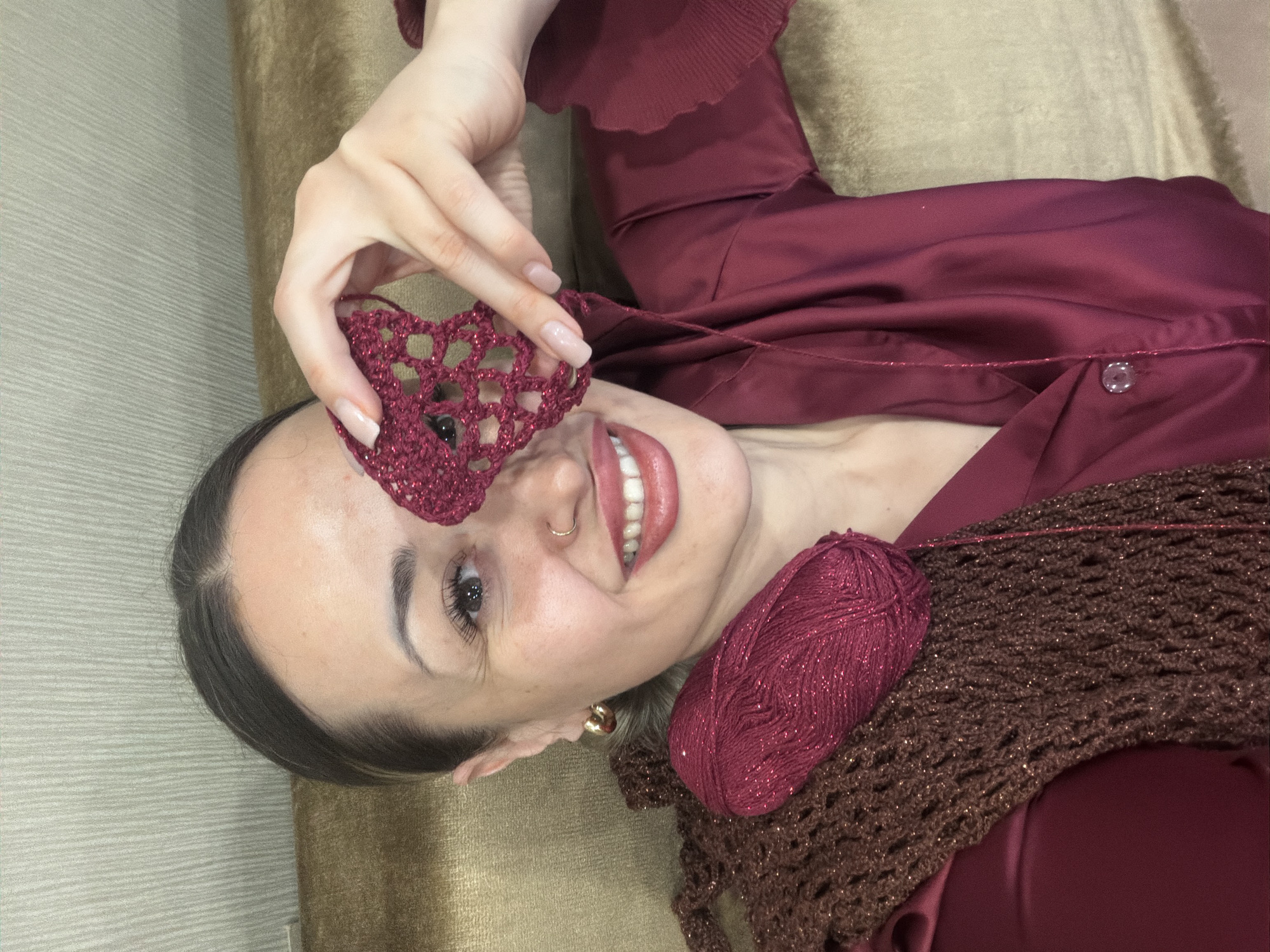 A woman is lying on a brown surface. She is wearing a dark red satin-like shirt. She is holding a crocheted heart-shaped object in front of her left eye. A ball of red yarn sits on top of a crocheted brown shawl or garment next to her. She is smiling. A gold-colored nose ring is visible. The location cannot be determined from the visual data.
