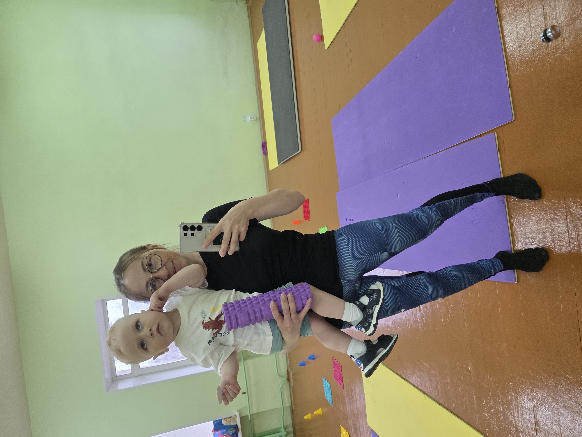This image captures a mirror selfie featuring a woman and a young child within an indoor setting, likely a gymnasium, physical therapy room, or activity center. The woman, with blonde hair and glasses, is dressed in a black t-shirt and blue patterned leggings. She holds a silver smartphone, reflecting her face with a subtle smile. She is holding a light-haired, blue-eyed toddler, who wears a white t-shirt with an unclear graphic or foreign text, light-colored shorts, and black and white sandals. The child is holding a purple textured foam roller.
The room features a light green wall on one side and a brown/orange wall and a shiny matching floor on the other. Natural light streams in from a window positioned behind the child. Various colored exercise or play mats, including purple, gray, and yellow, are laid out on the floor. Scattered around are small colorful cones (blue, orange, yellow, pink) and a pink ball, suggesting equipment for play, exercise, or therapy. Wall outlets are visible on the light green wall.
The scene depicts a casual, positive interaction between the woman and child, likely during a session or playtime focused on physical activity, given the equipment present. The clear natural light indicates it is daytime.