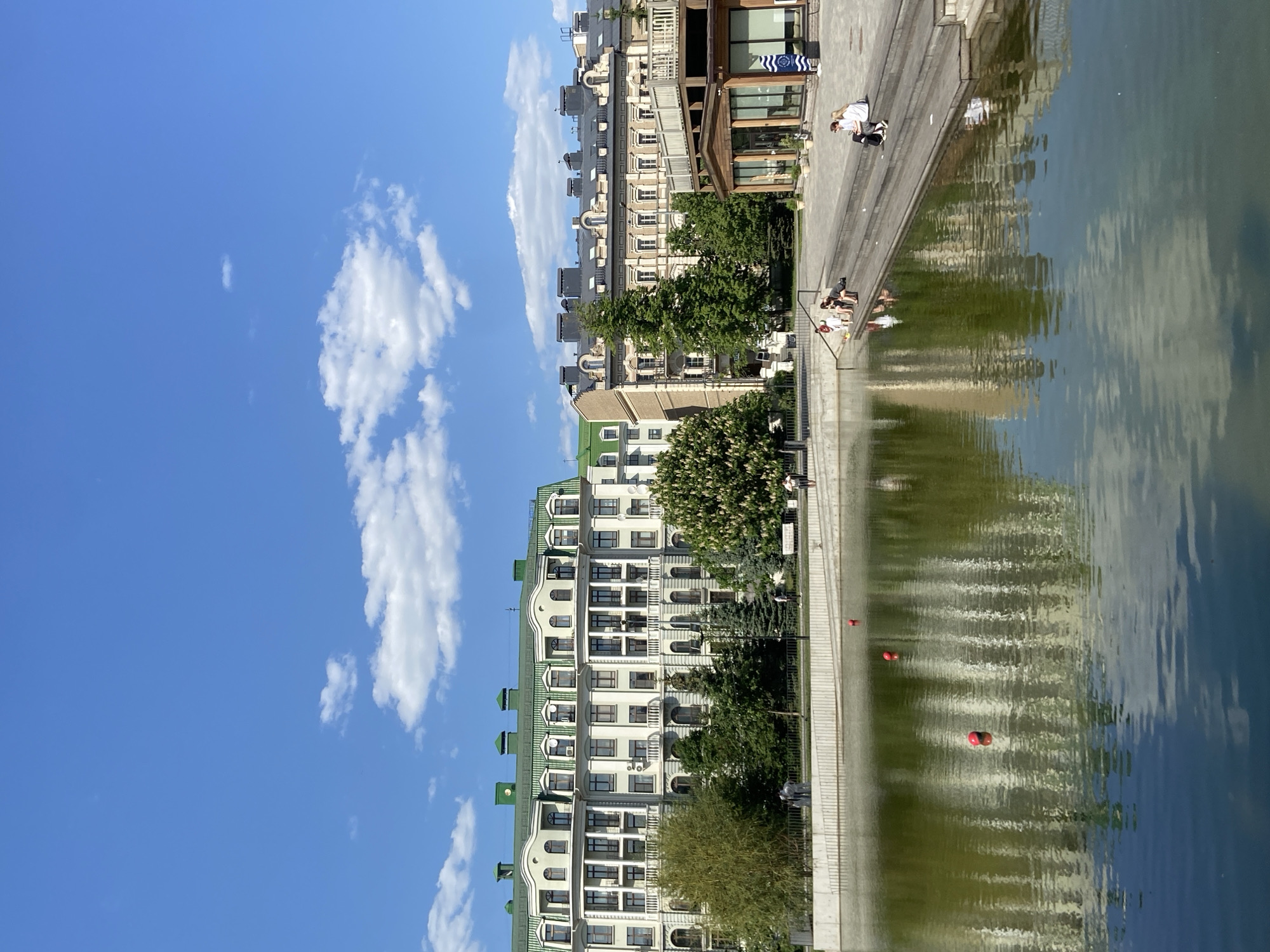 The image depicts a waterfront scene in Kazan, Russia. A multi-story building with a green roofline stands adjacent to a body of water reflecting the sky and adjacent structures. A pedestrian promenade lines the water's edge, where several people are seated or standing. Red buoys are visible in the water. The sky is blue with scattered white clouds.
