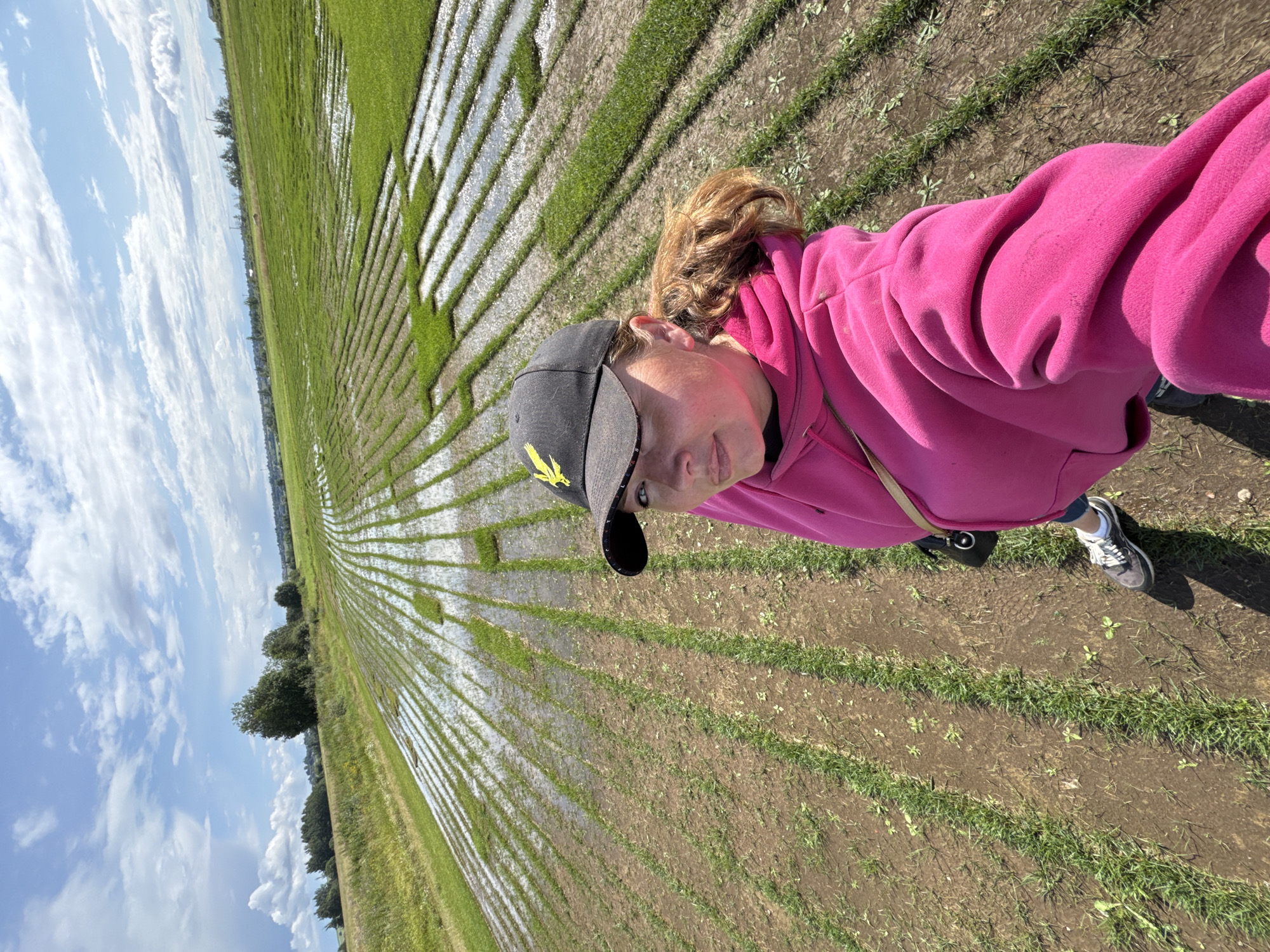 A woman in a bright pink hoodie and a black baseball cap with a yellow emblem is taking a selfie in a flooded field. The field is divided into parallel rows, with water pooled in some of them, creating a striped effect. The woman's hair is pulled back into a ponytail. In the background, there are trees and a cloudy sky. The scene appears to be in a rural area.